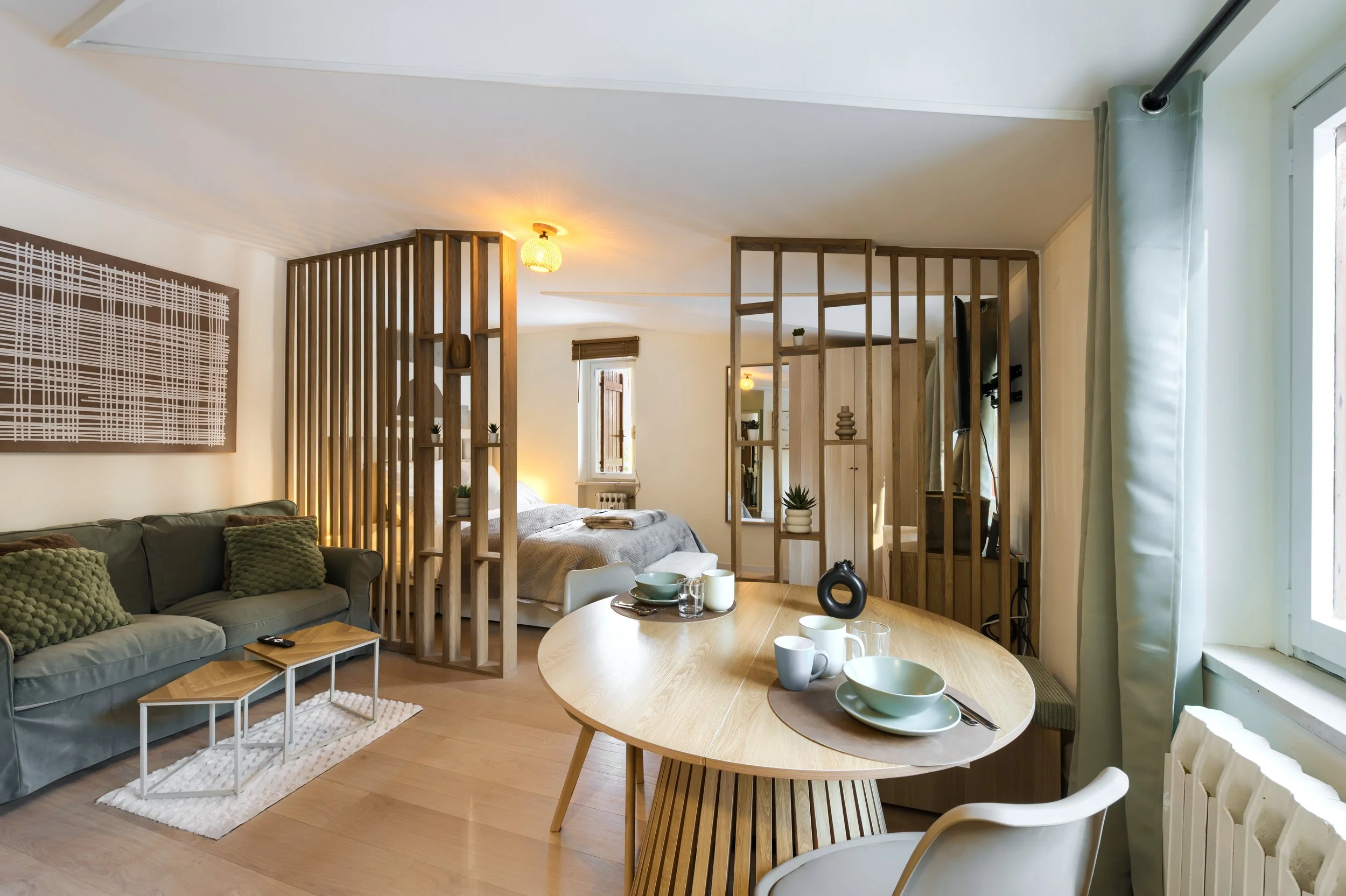 Open-plan living and sleeping area with a wooden partition, a sofa with green cushions, a round wooden dining table with dishes, and a bed with gray bedding, natural light from windows, a luxurious retreat, an exclusive retreat.