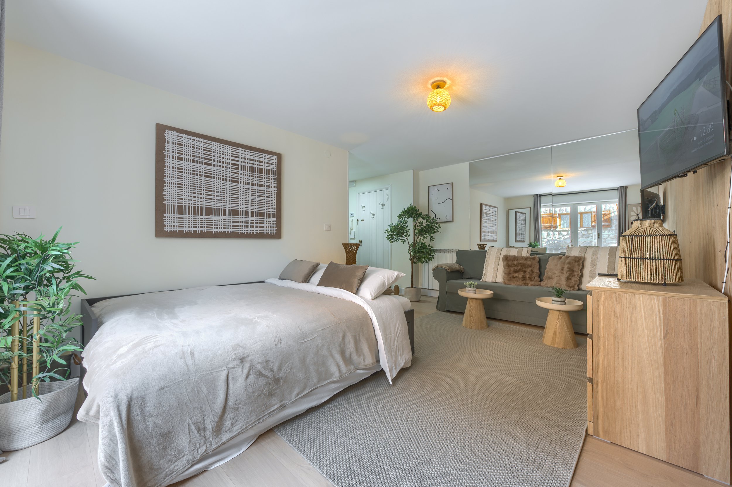 A cozy living room with a bed on the left, decorated with pillows, and a green sofa with fur cushions on the right. There are small round tables, a wooden cabinet with a lamp, a flat-screen TV on the wall, and large windows with curtains allowing nat