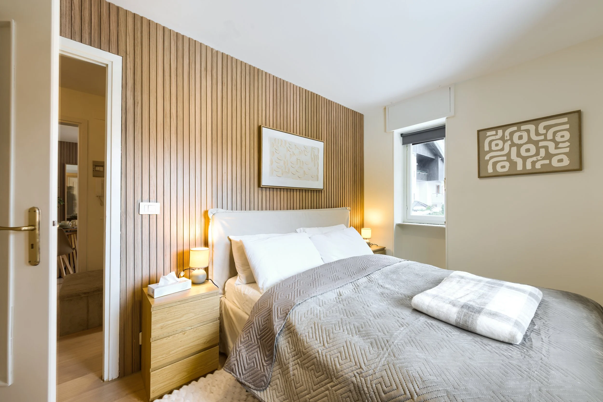 A modern bedroom with a wooden accent wall behind a bed with white pillows and a beige quilted bedspread. There are two lamps on nightstands on either side of the bed and abstract artwork on the walls. a luxurious retreat, an exclusive retreat.