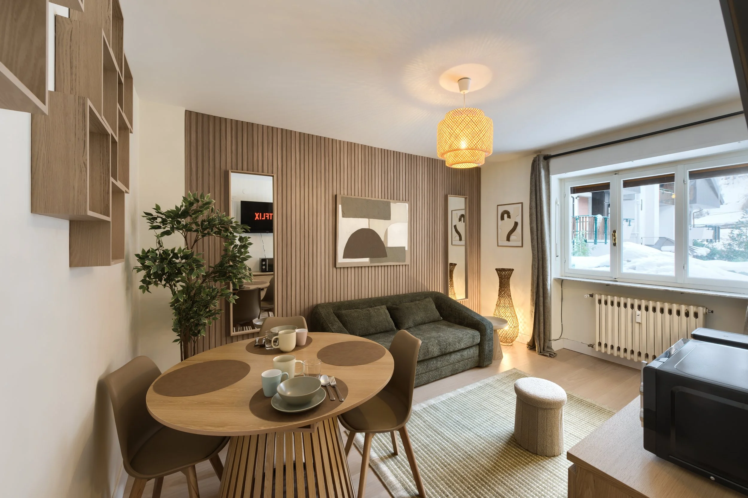 Living room and dining area with a round wooden table set with cups and plates, a green sofa, wall art, large window with a snow-covered exterior, a TV, and a potted plant, a luxurious retreat, an exclusive retreat, a refined mountain retreat.