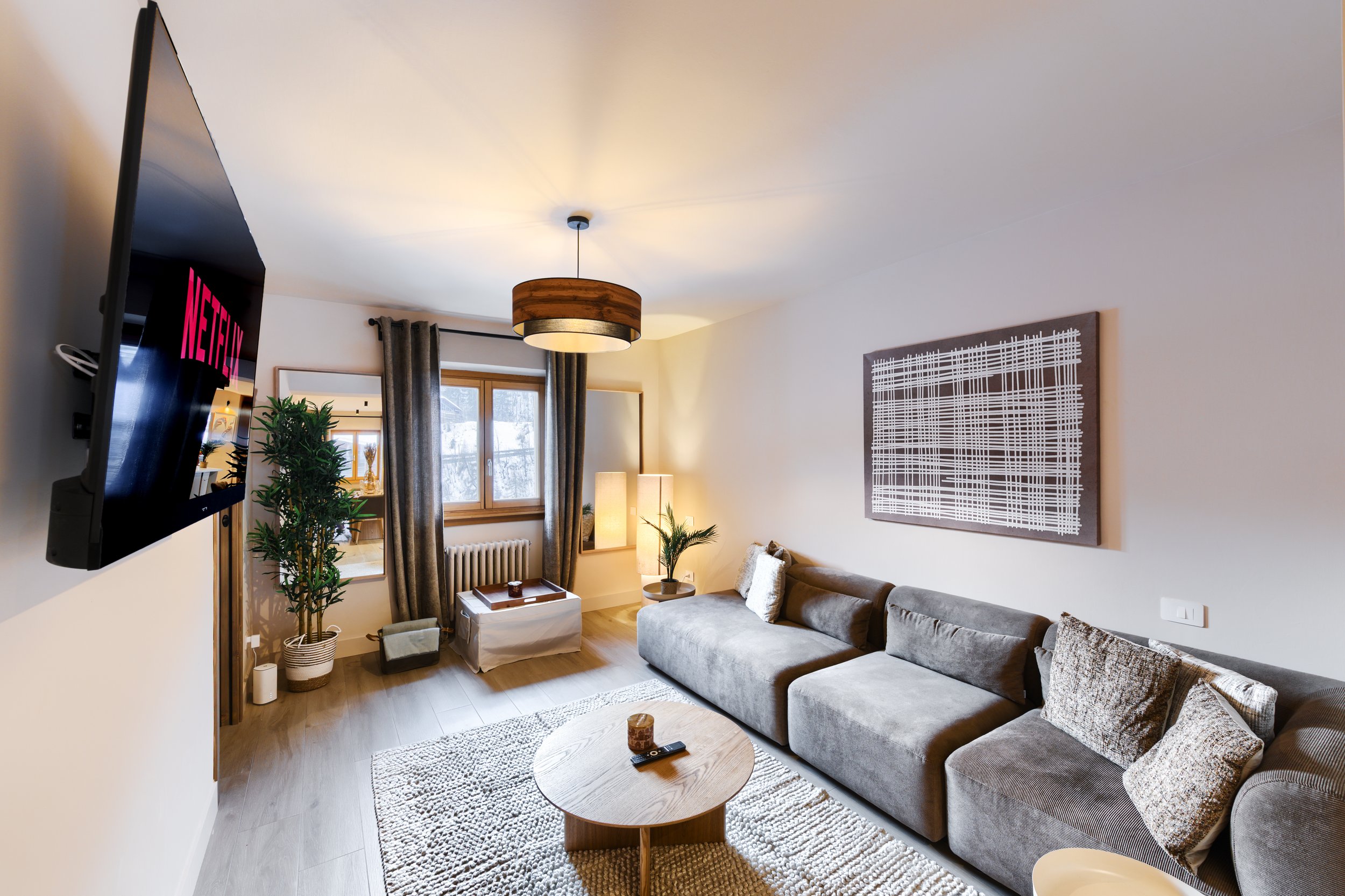 Living room with a gray sectional sofa, a small round wooden coffee table, a striped rug, potted plants, window with curtains, a wall-mounted TV, and wall art, a luxurious retreat, an exclusive retreat, a refined mountain retreat, premium alpine.
