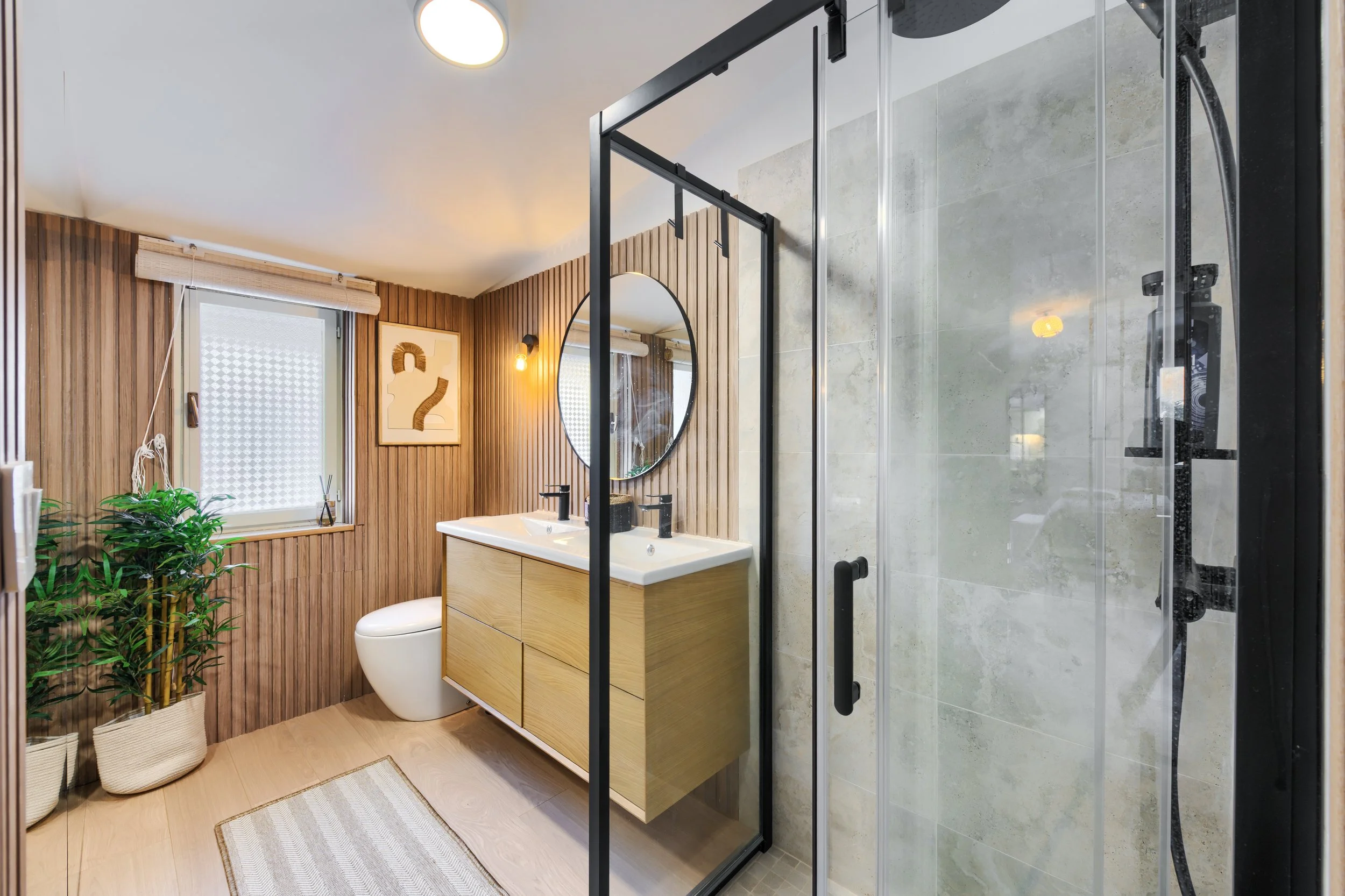 Modern bathroom with wooden wall paneling, a double sink vanity with black fixtures, a round mirror, a framed art piece, a window with a blind, a potted plant, and a glass shower enclosure with black hardware, a luxurious retreat, exclusive retreat.