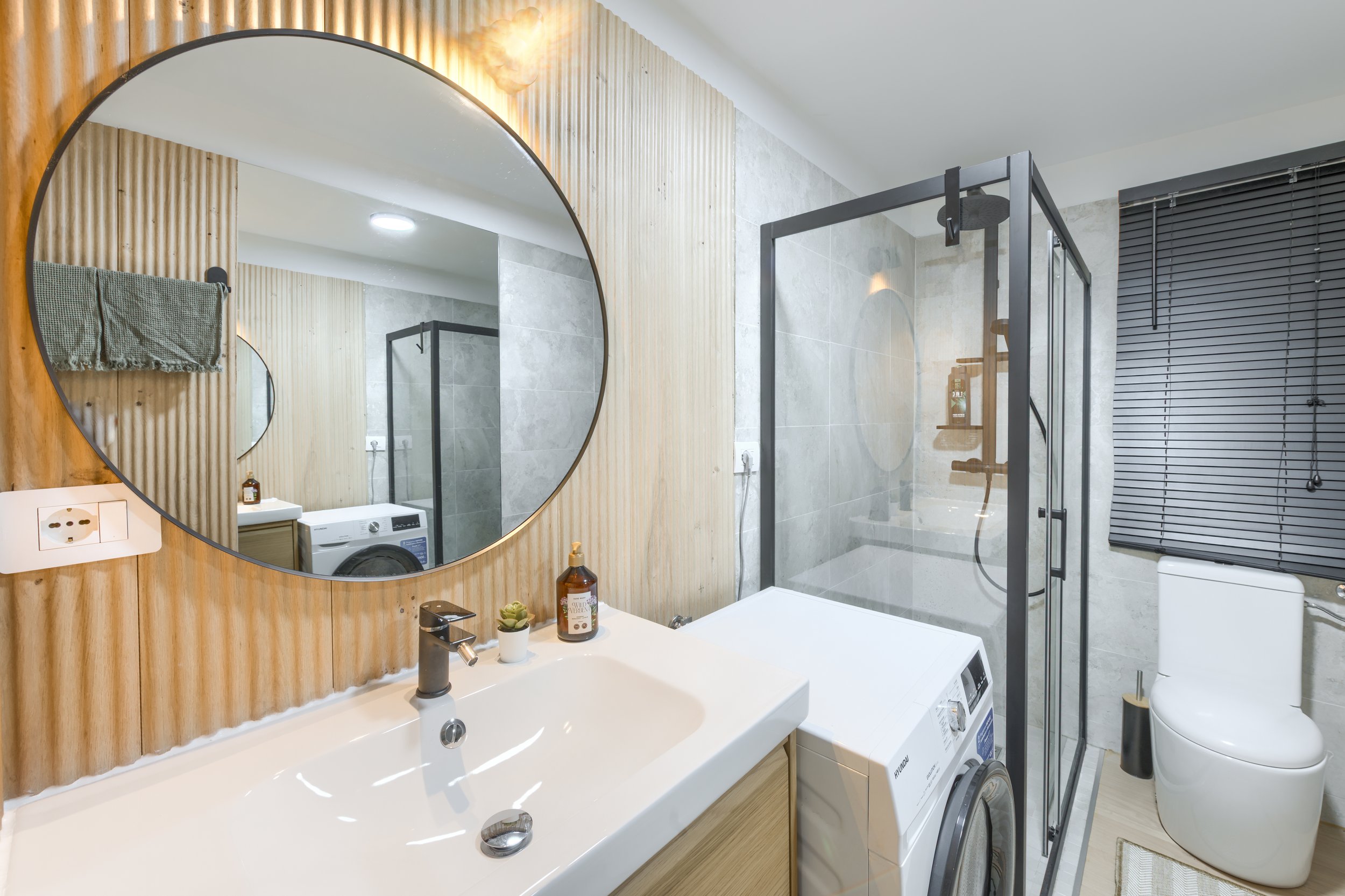 Modern bathroom with large round mirror, wooden wall paneling, white sink, bottle of soap, small plant, washing machine, glass shower enclosure, toilet, and window with black blinds, a luxurious retreat, an exclusive retreat, refined mountain retreat