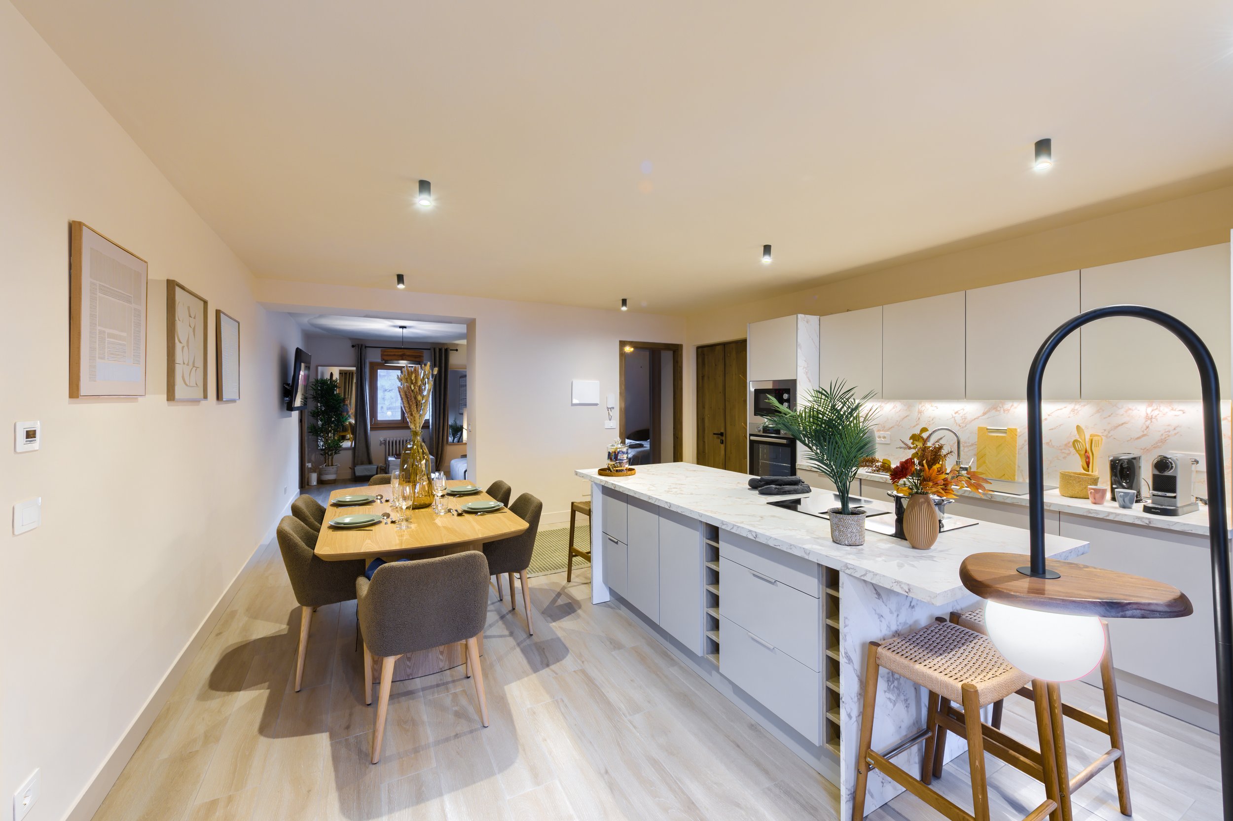 Modern open-concept kitchen and dining area with white cabinetry, marble island, wooden dining table, and beige chairs, decorated with plants and framed artwork, a luxurious retreat, an exclusive retreat, a refined mountain retreat, premium alpine. 