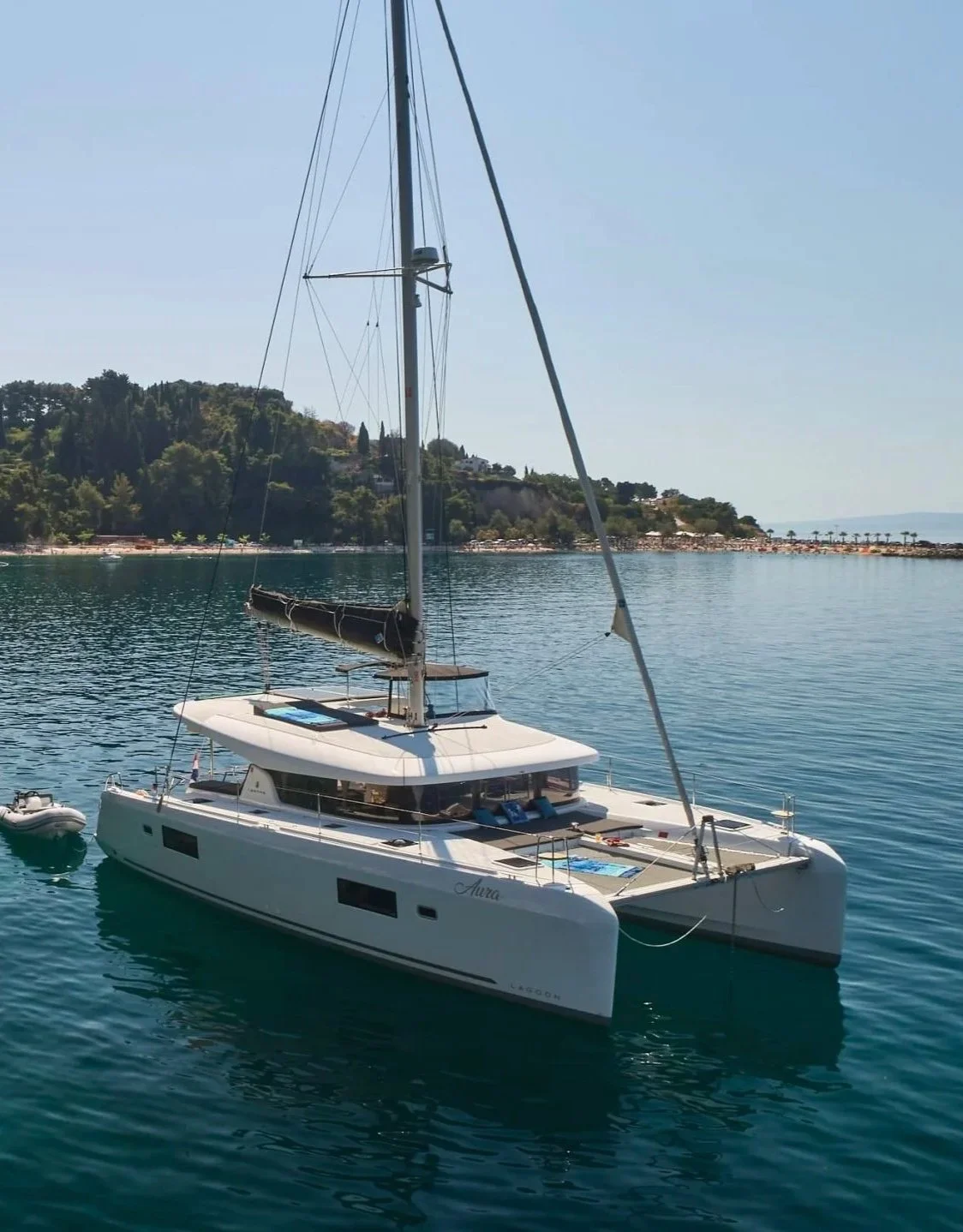 Catamaran Aura with black sails in a calm body of water, with trees and hills in the background under a clear blue sky.