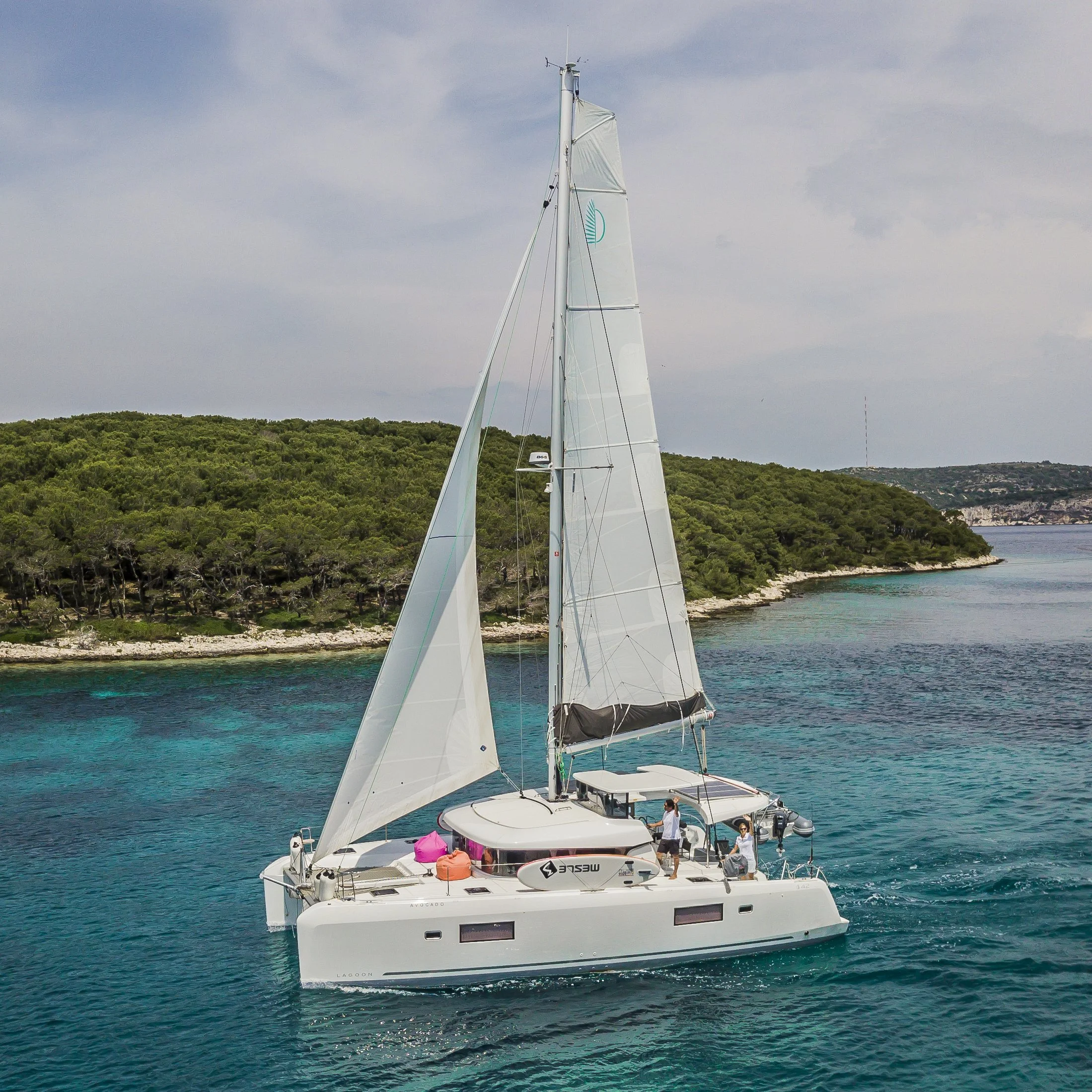 A white sailing catamaran with two tall masts and sails, floating on clear blue water near a green, hilly coastline under a partly cloudy sky.