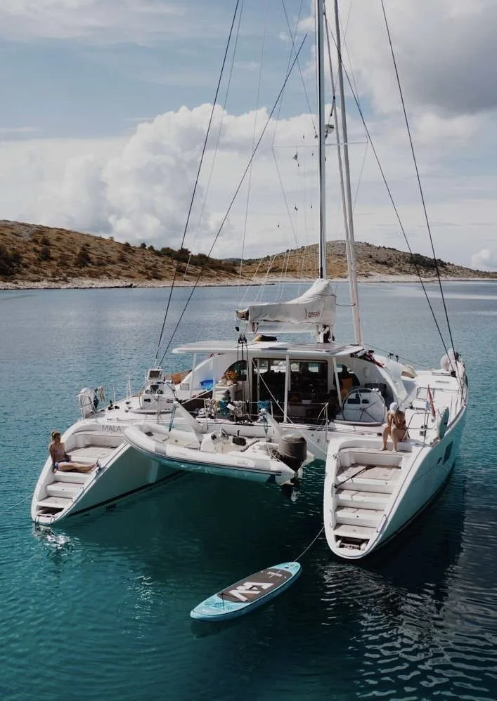 Catamaran MALA anchored in a calm turquoise body of water with two people relaxing on the deck. One person is sitting near the steps on the left side, and another is kneeling on the right side. A paddleboard is floating nearby.