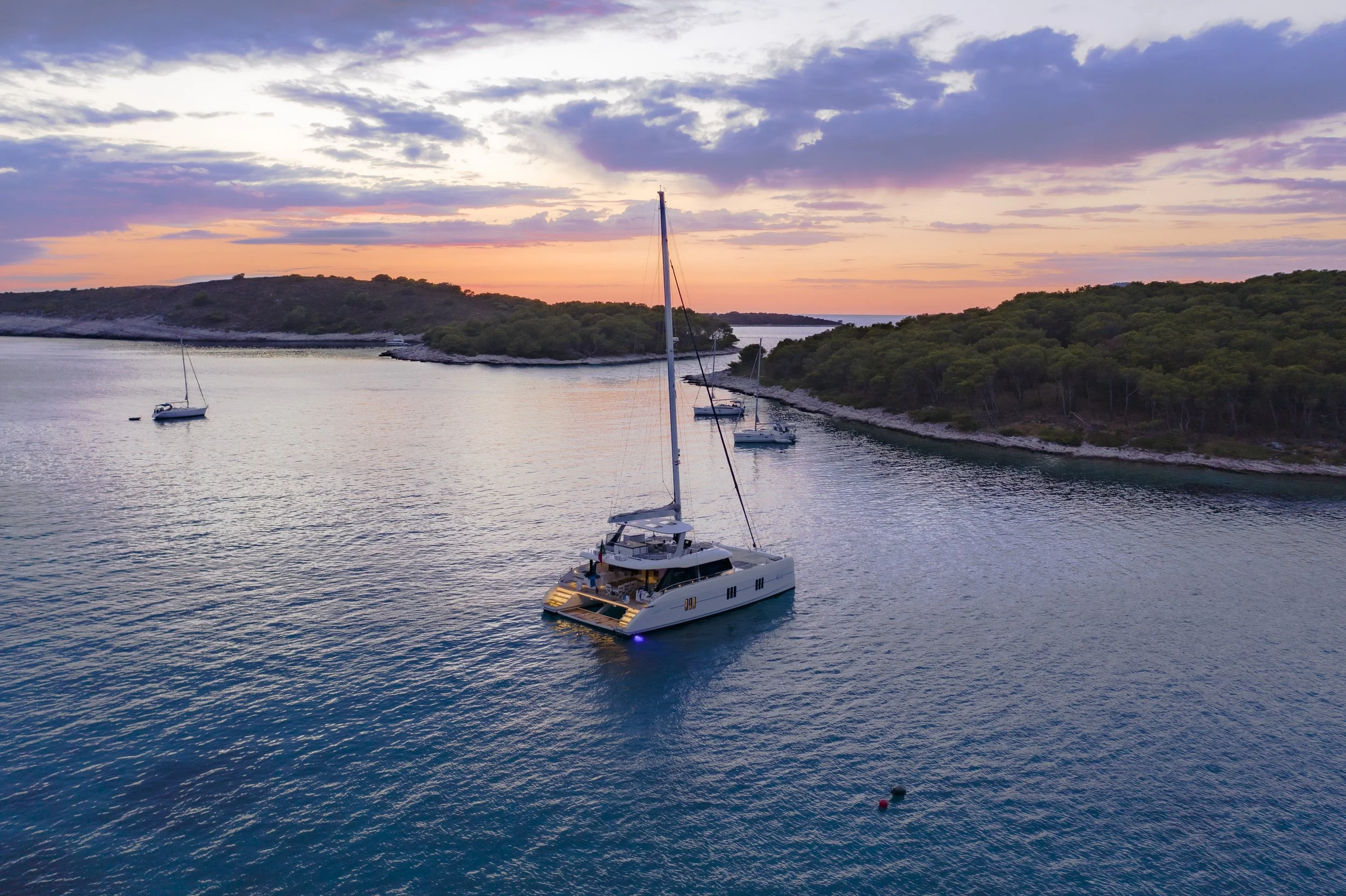 Sailboats anchored in a calm bay during sunset, with lush green islands and a partly cloudy sky in the background.