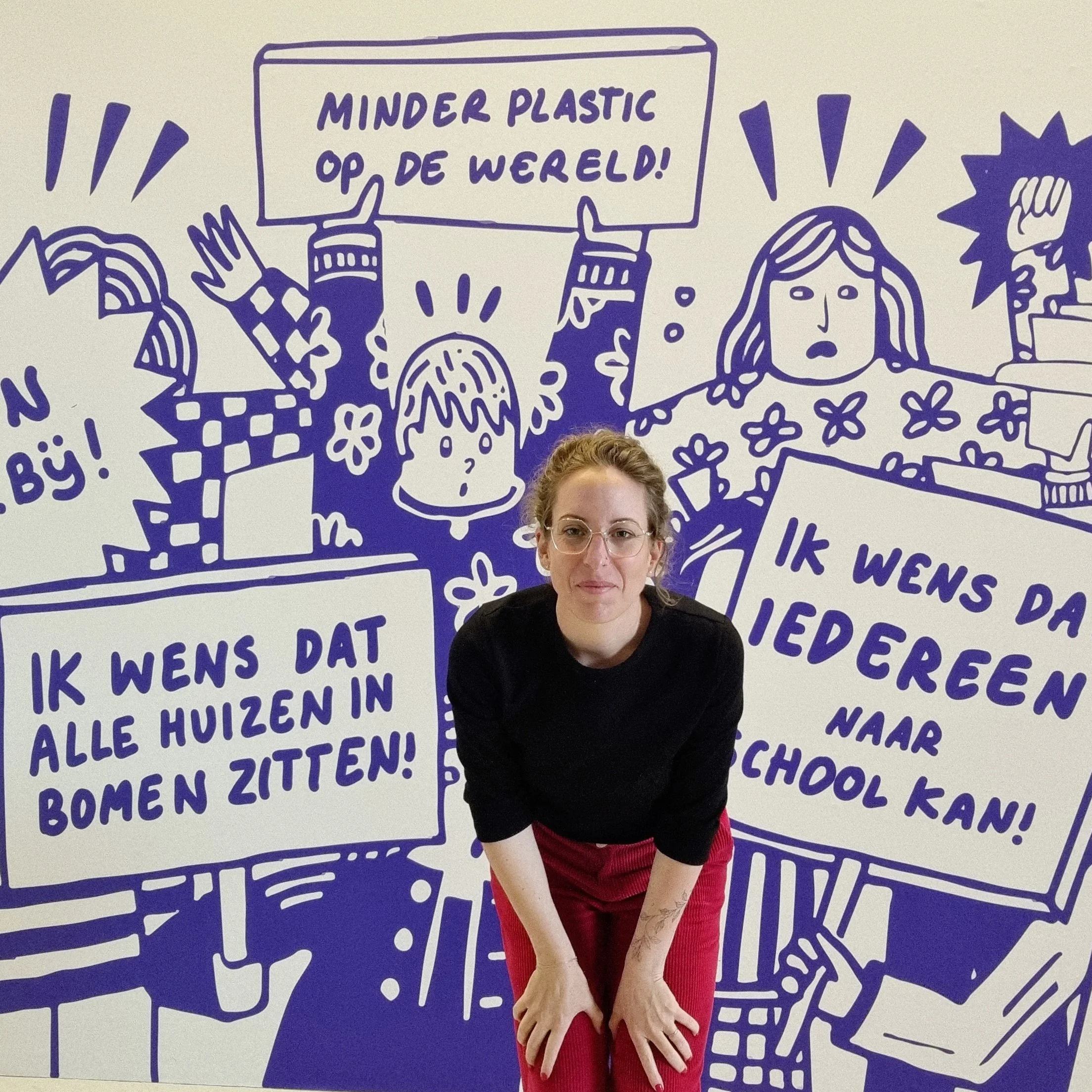 A woman with glasses and curly hair posing in front of a mural with cartoons and speech bubbles in Dutch.