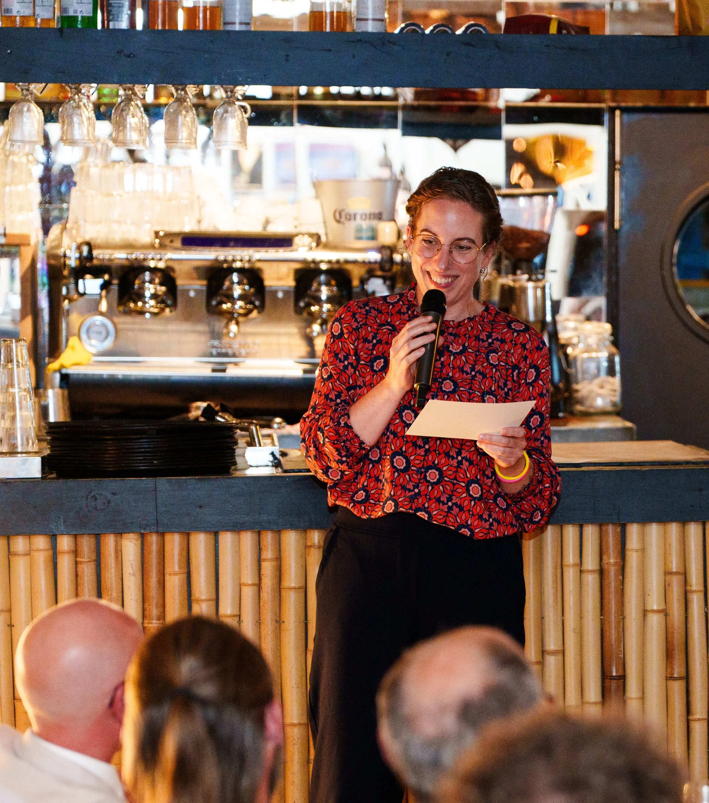 A woman in a red patterned blouse holding a microphone and reading from a paper, standing in front of a bar with coffee equipment, glasses, and bottles. She is smiling and speaking to an audience.
