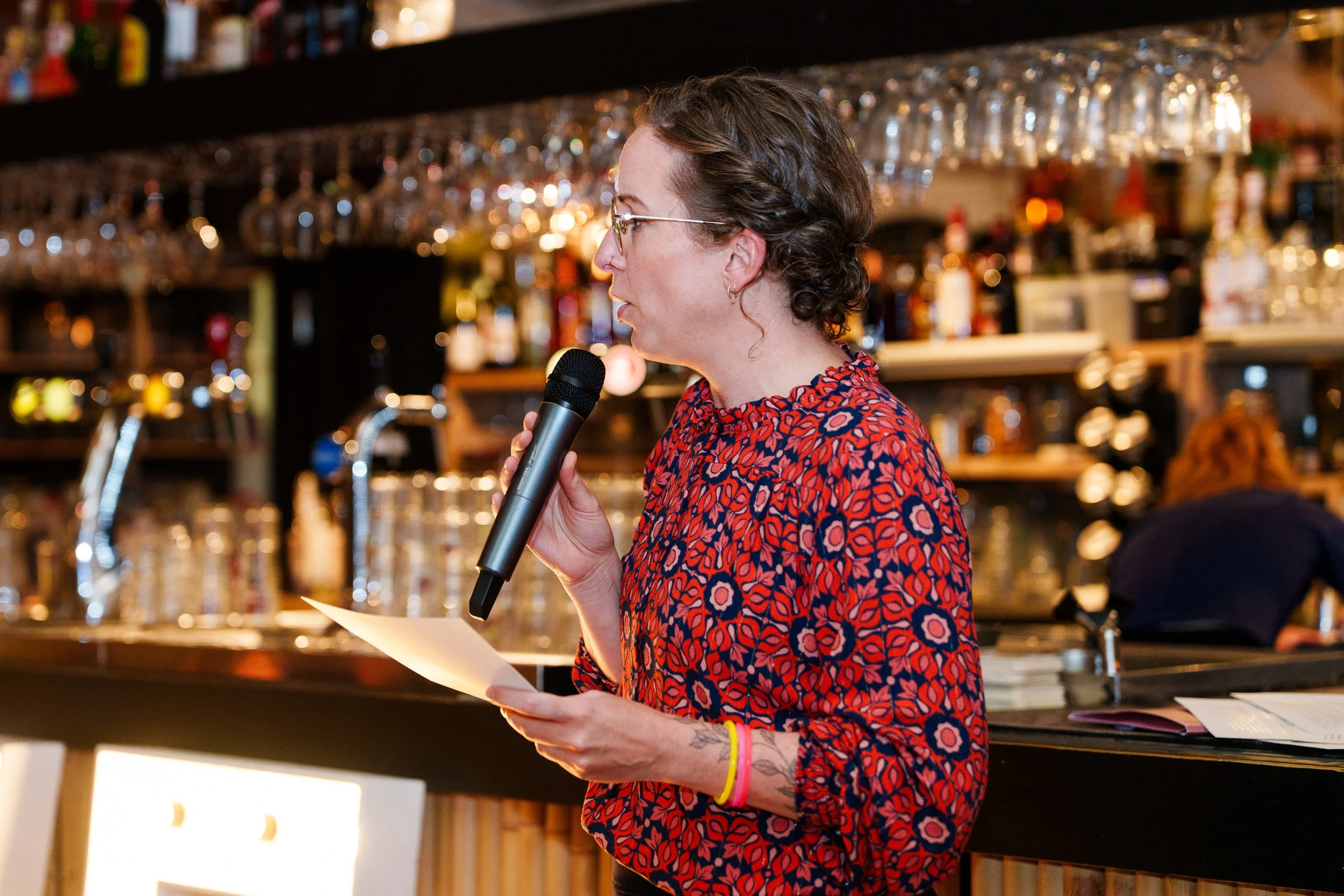 A woman with short curly hair and glasses holding a microphone and reading from a piece of paper in a bar or restaurant setting, with shelves of bottles and glasses in the background.