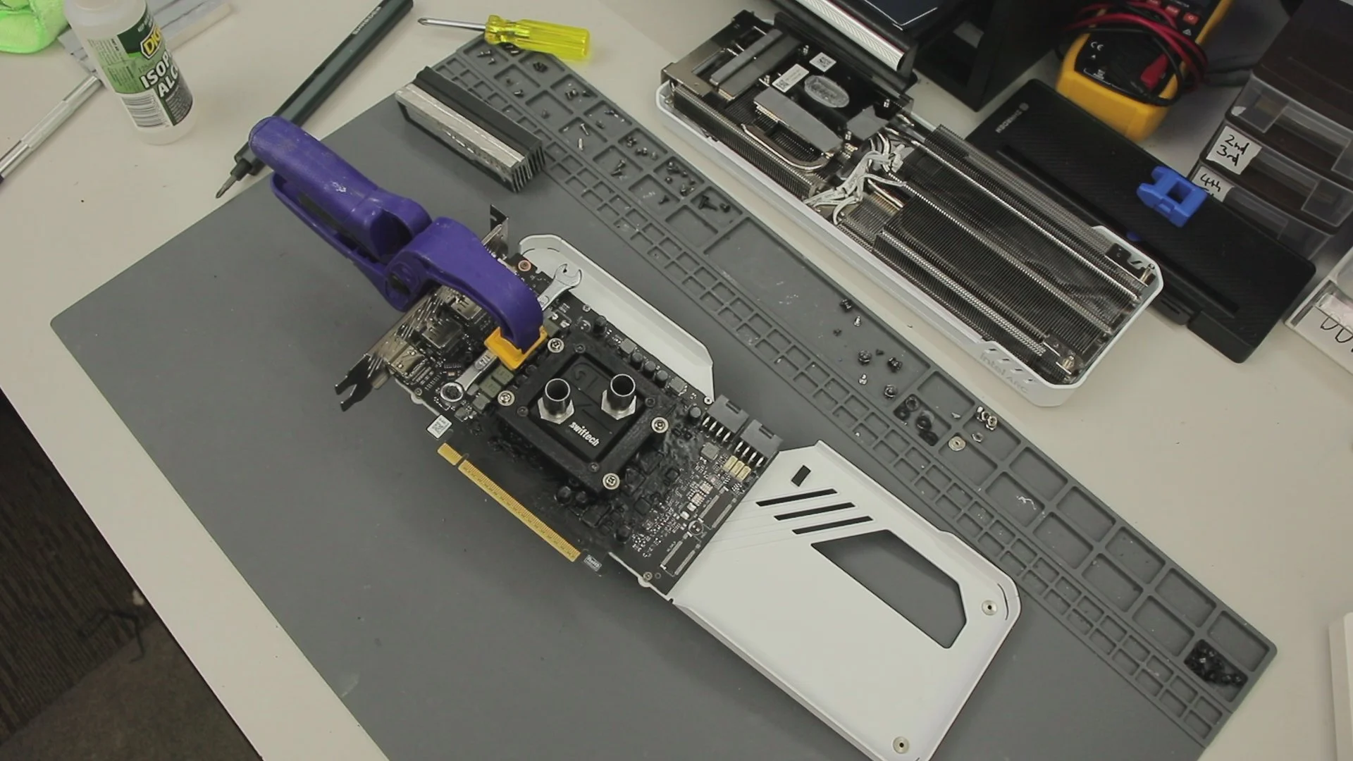 Computer graphics card with a liquid cooling block attached, on a work table with tools and disassembled computer parts.