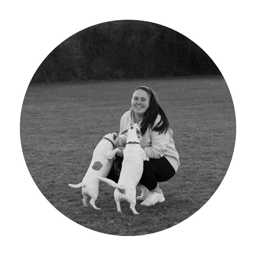 A woman smiling while squatting in a grassy field with two dogs that are standing on hind legs, one with a spots on its side, in a circular crop, black and white photo.