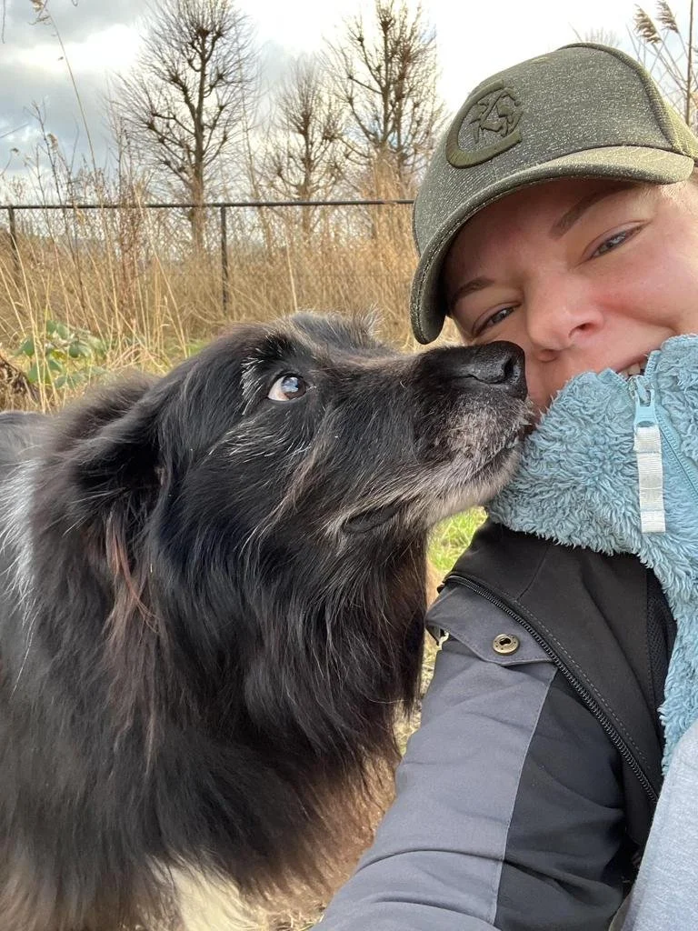 Een persoon met een pet die knuffelt met een zwarte, pluizige hond in een outdoor omgeving met bomen en gras.