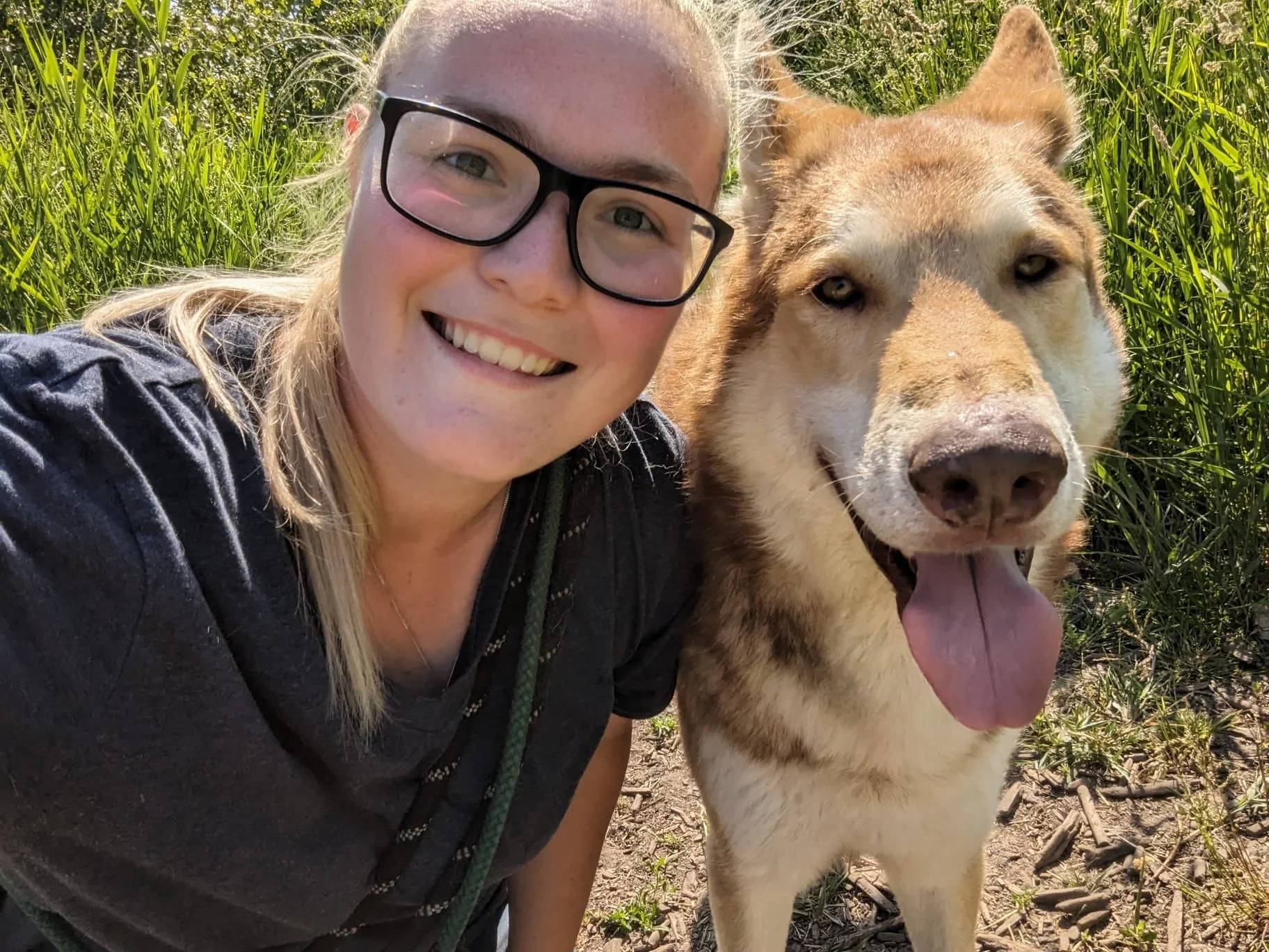Een glimlachende vrouw met zwarte bril poseert naast een grote, vriendelijke hond met een tong uit de mond in een groen veld.