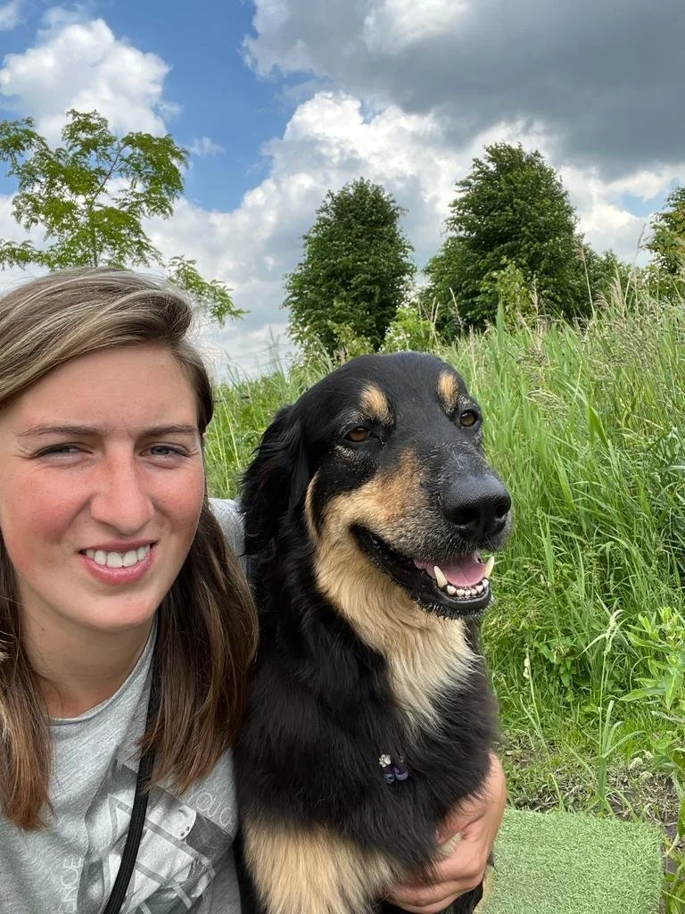 Een vrouw en haar hond die samen in een groene veld staan, met bomen en een bewolkte hemel op de achtergrond.