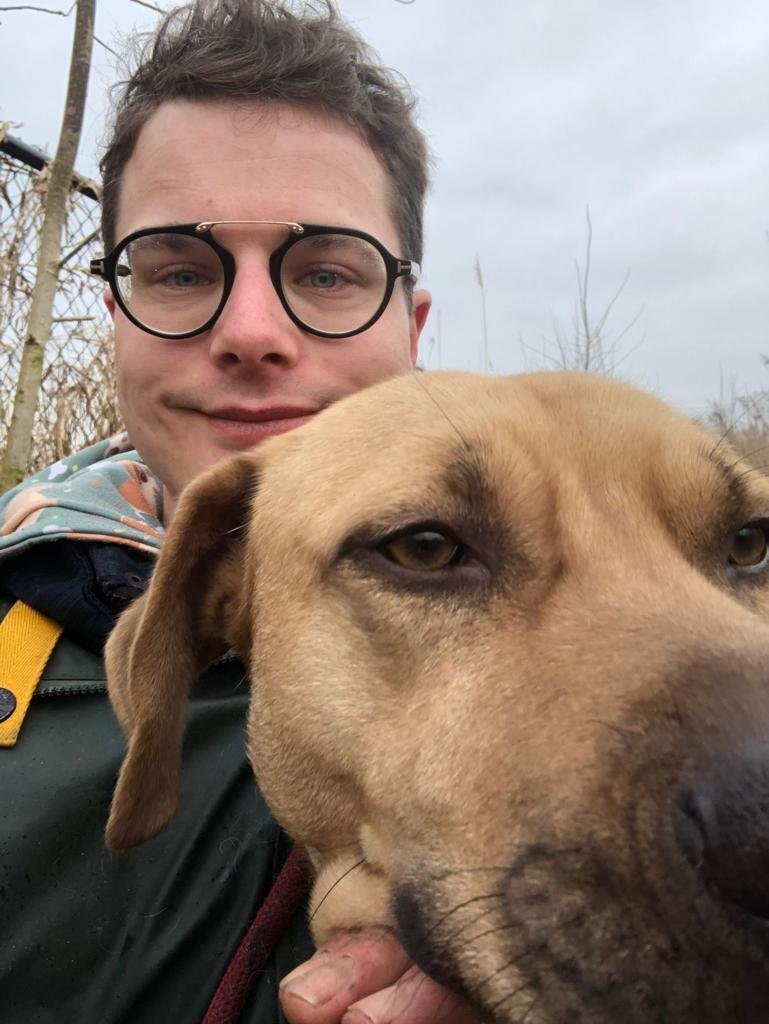 Een man met bril en een grote hond, mogelijk een bondgenootschap omringd door een natuurlijke omgeving met bomen en een bewolkte hemel.