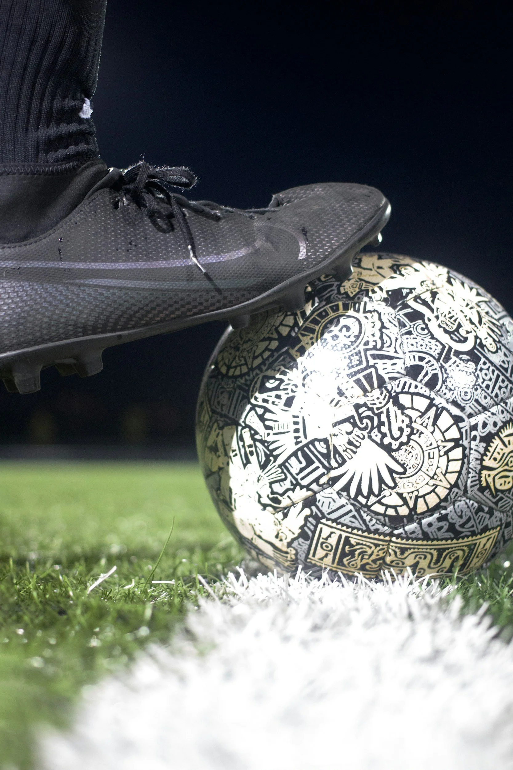 Close-up of a black football boot on a person's foot, balanced on a black and white patterned football on a pitch at night.