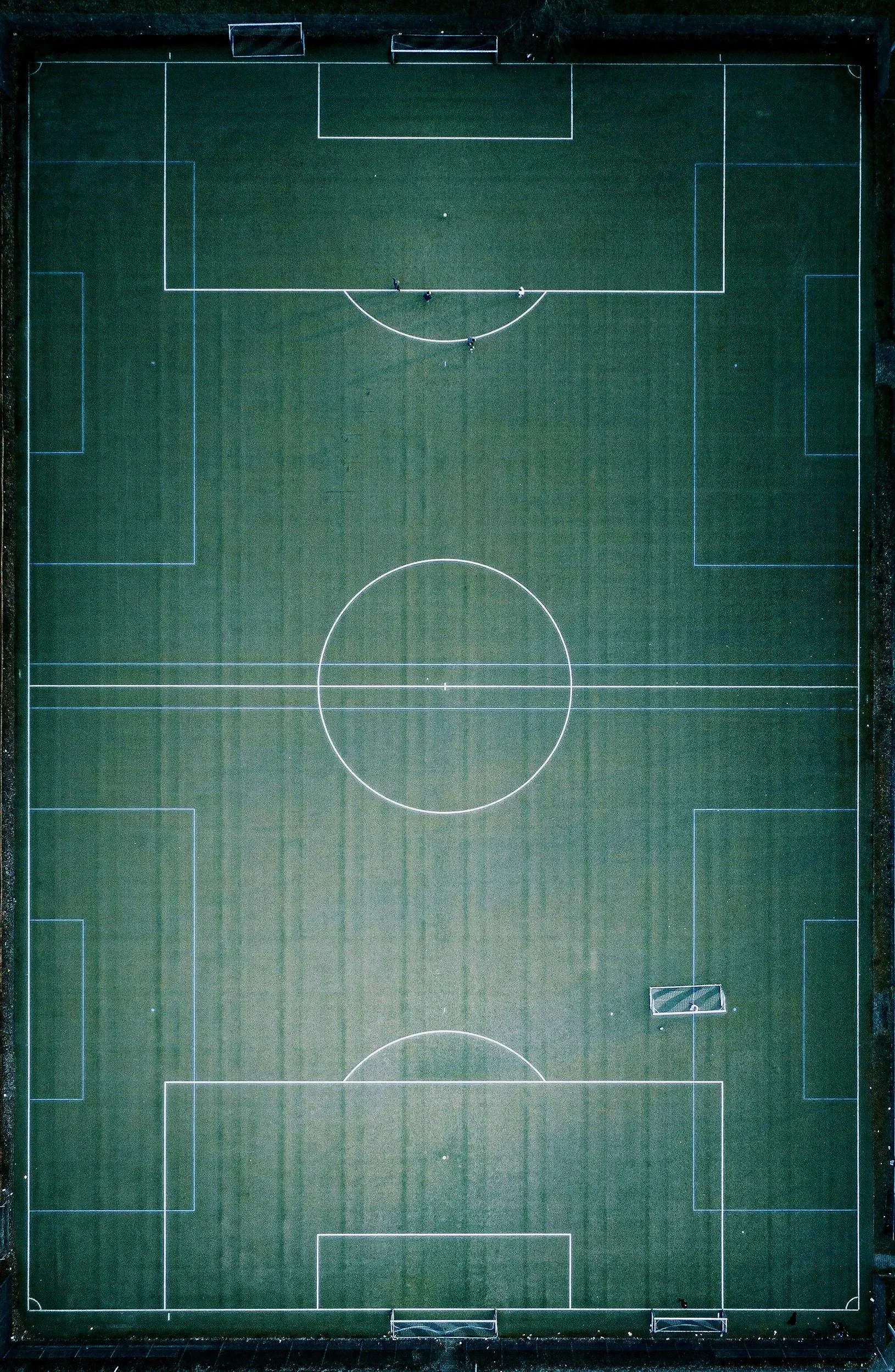 An aerial view of an empty outdoor football pitch with white boundary lines, including two goals, a centre circle, and penalty areas, surrounded by a dark perimeter.