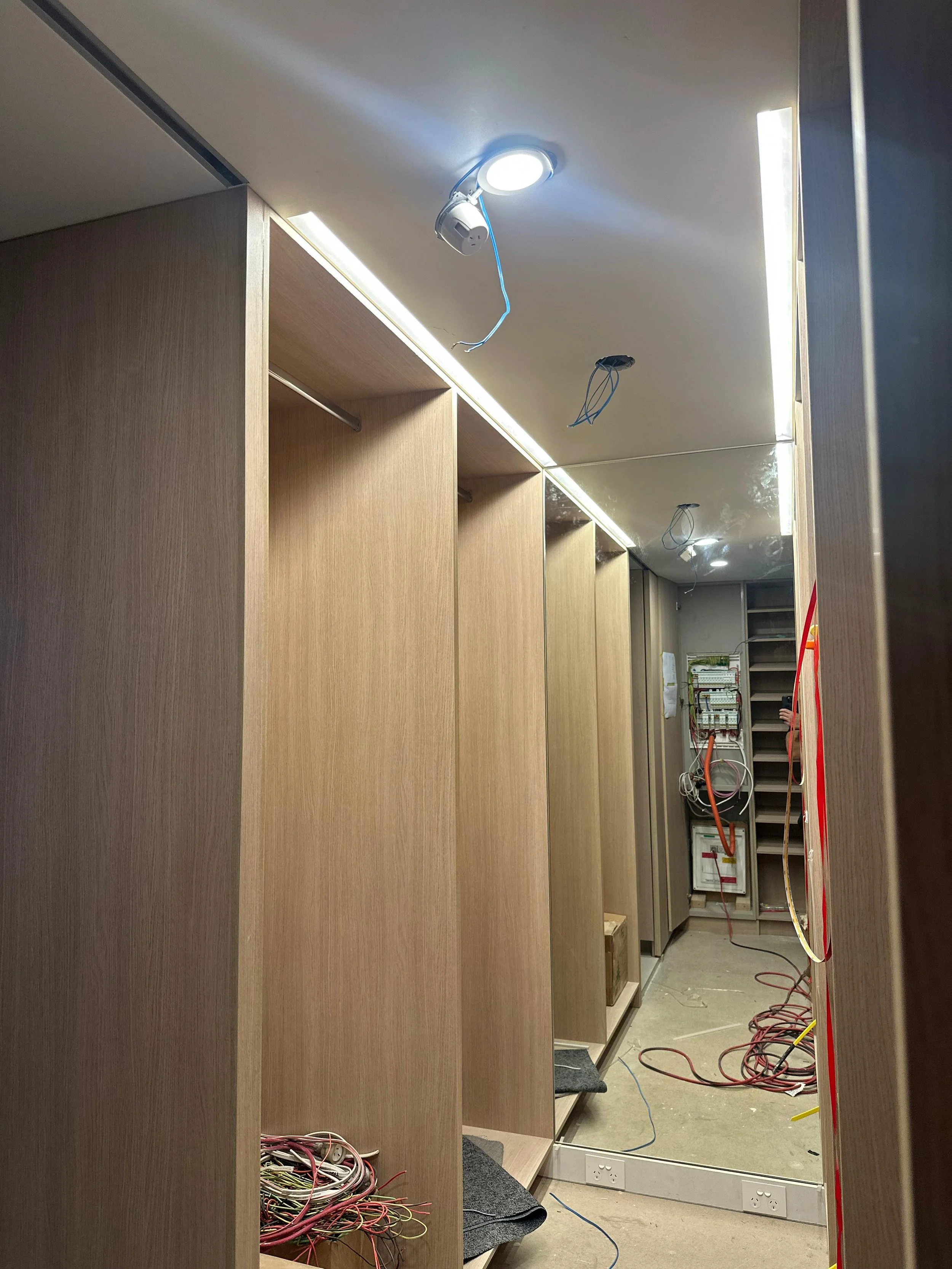 Interior view of a room under construction with wooden panels, electrical wires, and open electrical panels, ceiling lights, and a large mirror reflecting part of the room.