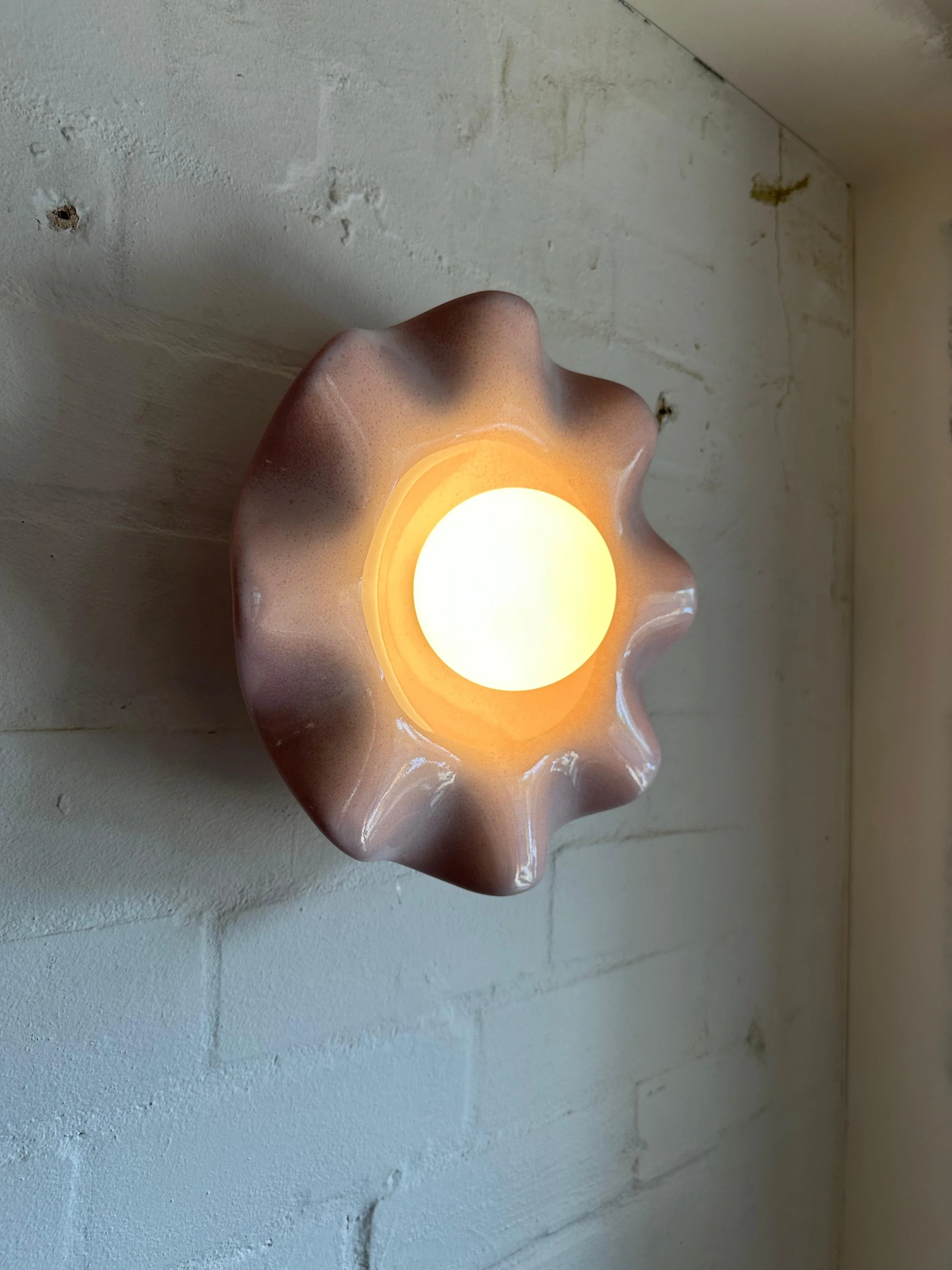 Wall-mounted lamp with a brown, wavy, petal-shaped ceramic or stone frame glowing with warm light, installed on a textured off-white brick wall.