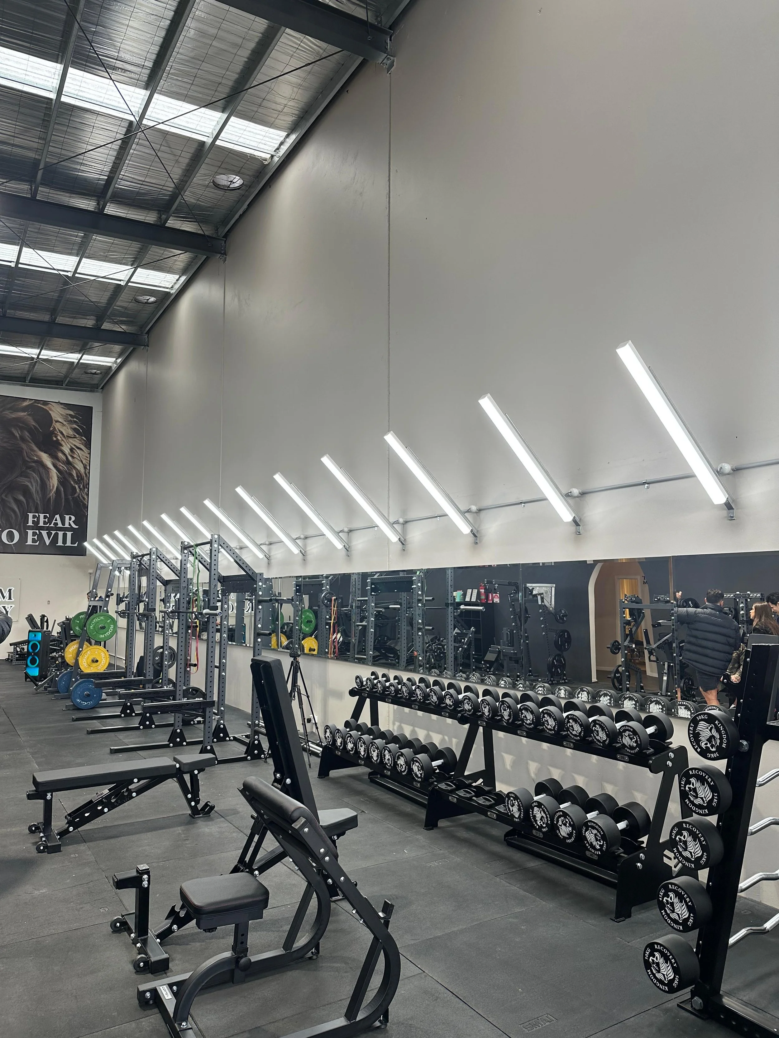 Interior of a gym with workout equipment including dumbbells, benches, and weight racks, illuminated by bright overhead lighting.