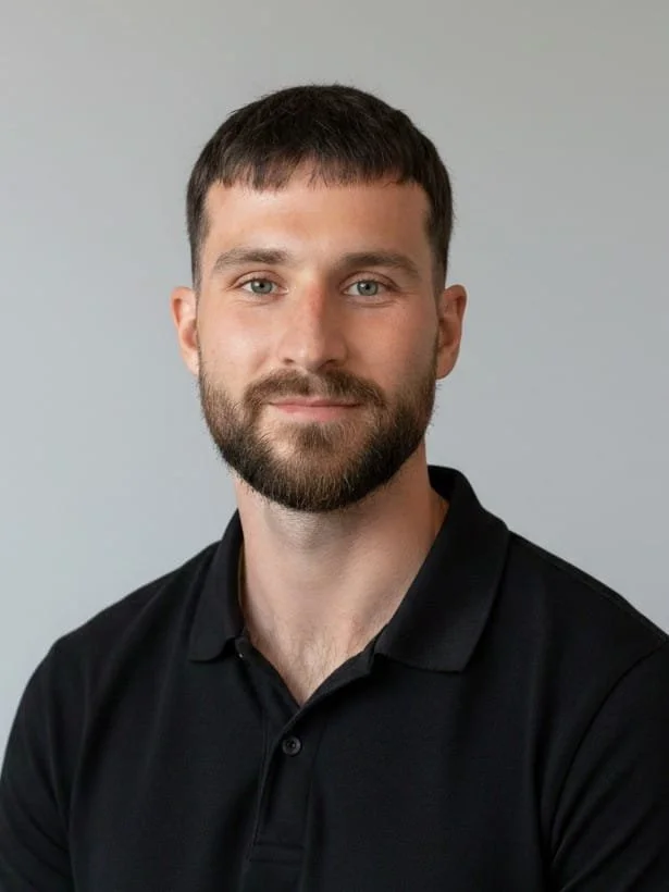 A man with short dark hair and a beard, wearing a black collared shirt, looking at the camera against a light gray background.