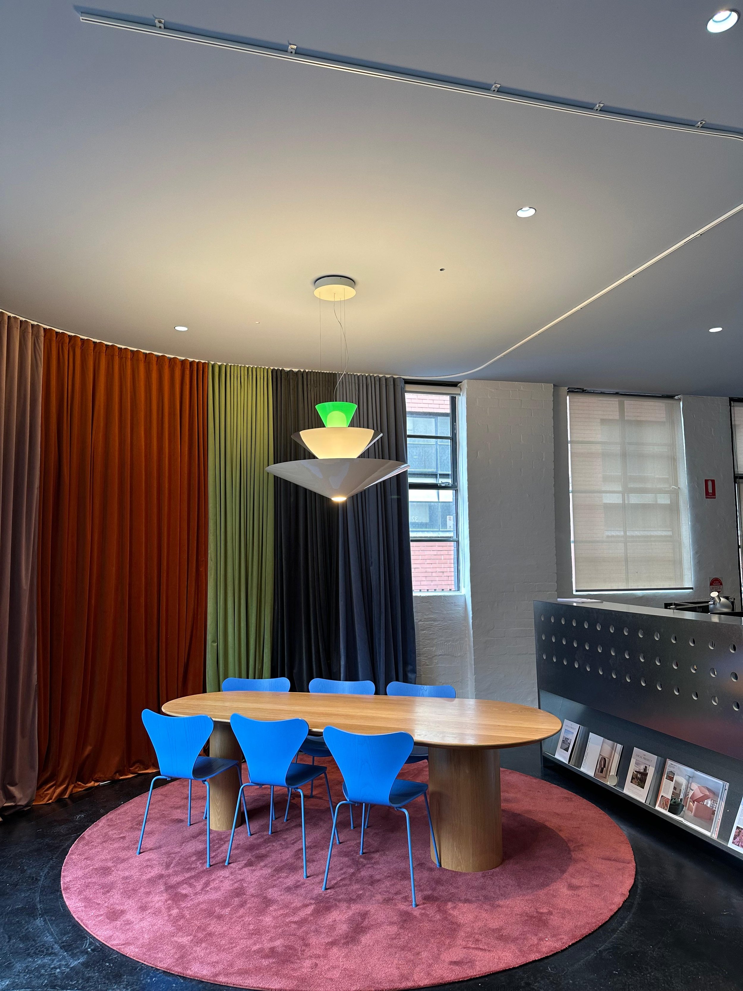 Interior of a modern room with a round wooden table, six blue chairs, a colorful rainbow curtain, a circular hanging light fixture, and large windows with blinds