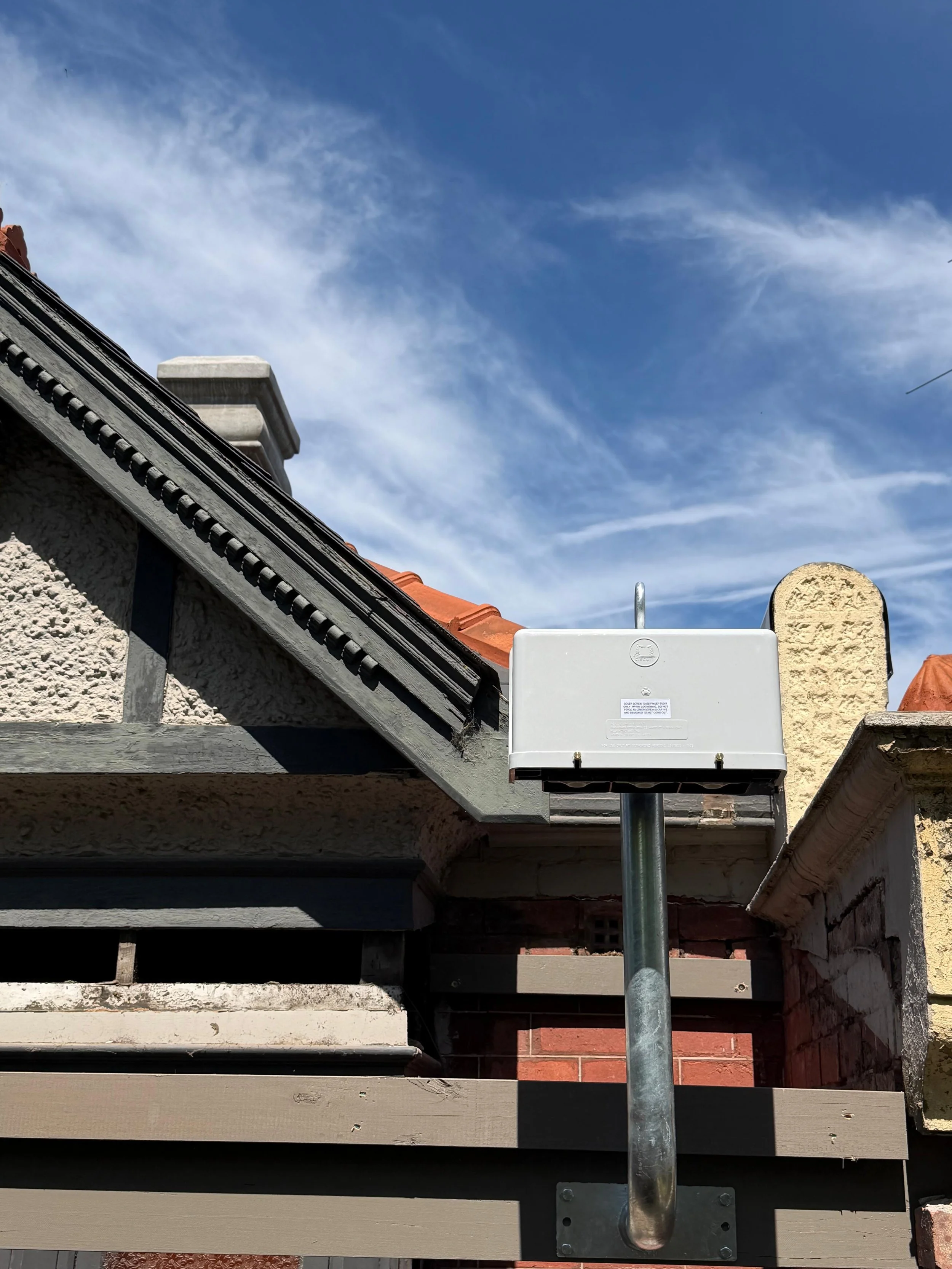 A weather sensor or antenna mounted on a metal pole on the roof of a house with a textured stucco wall, brick, and wood siding, under a blue sky with wispy clouds.