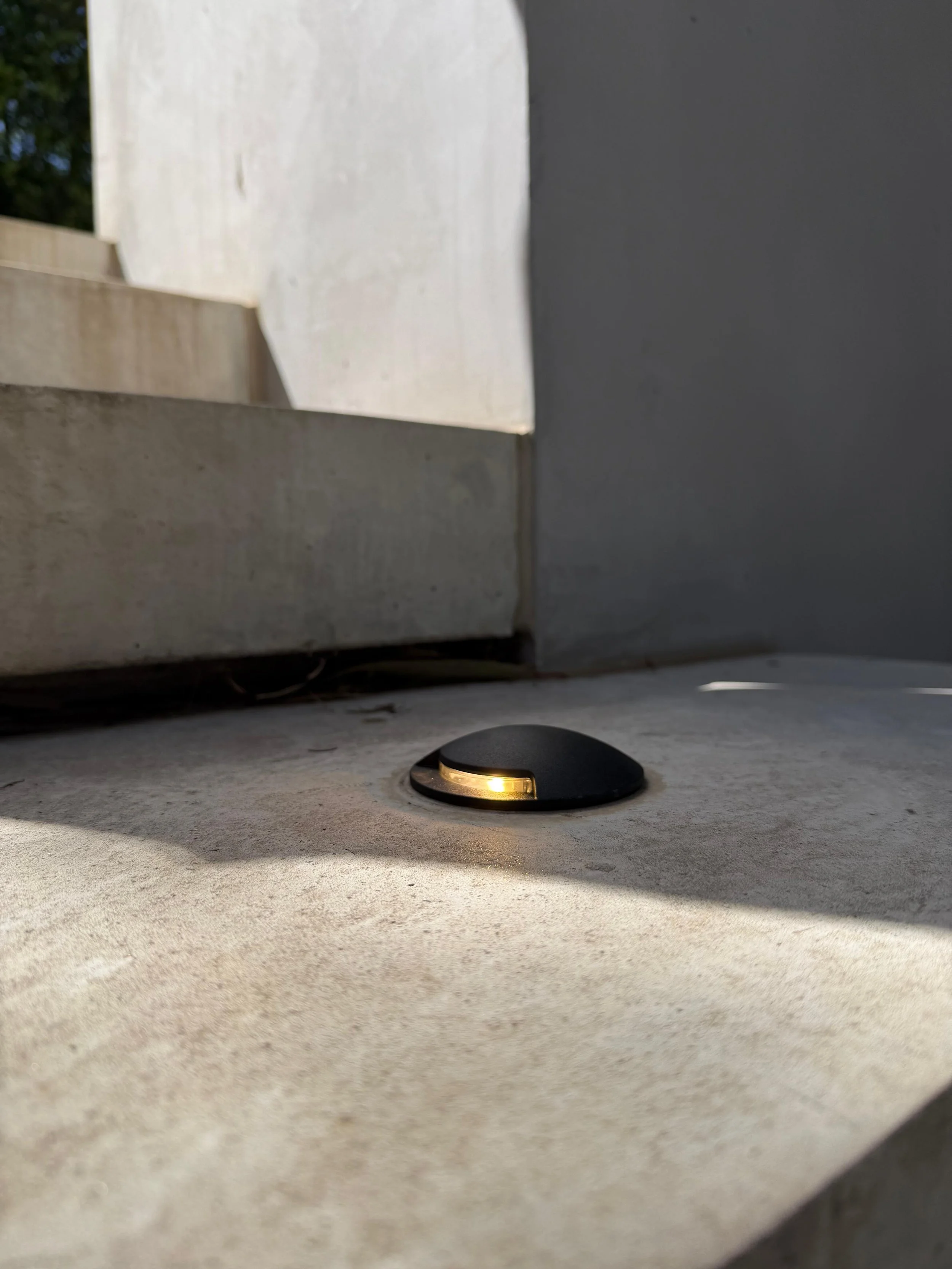 Close-up of a black, rounded outdoor light fixture with a small yellow light, installed at the base of a wall on a concrete surface.