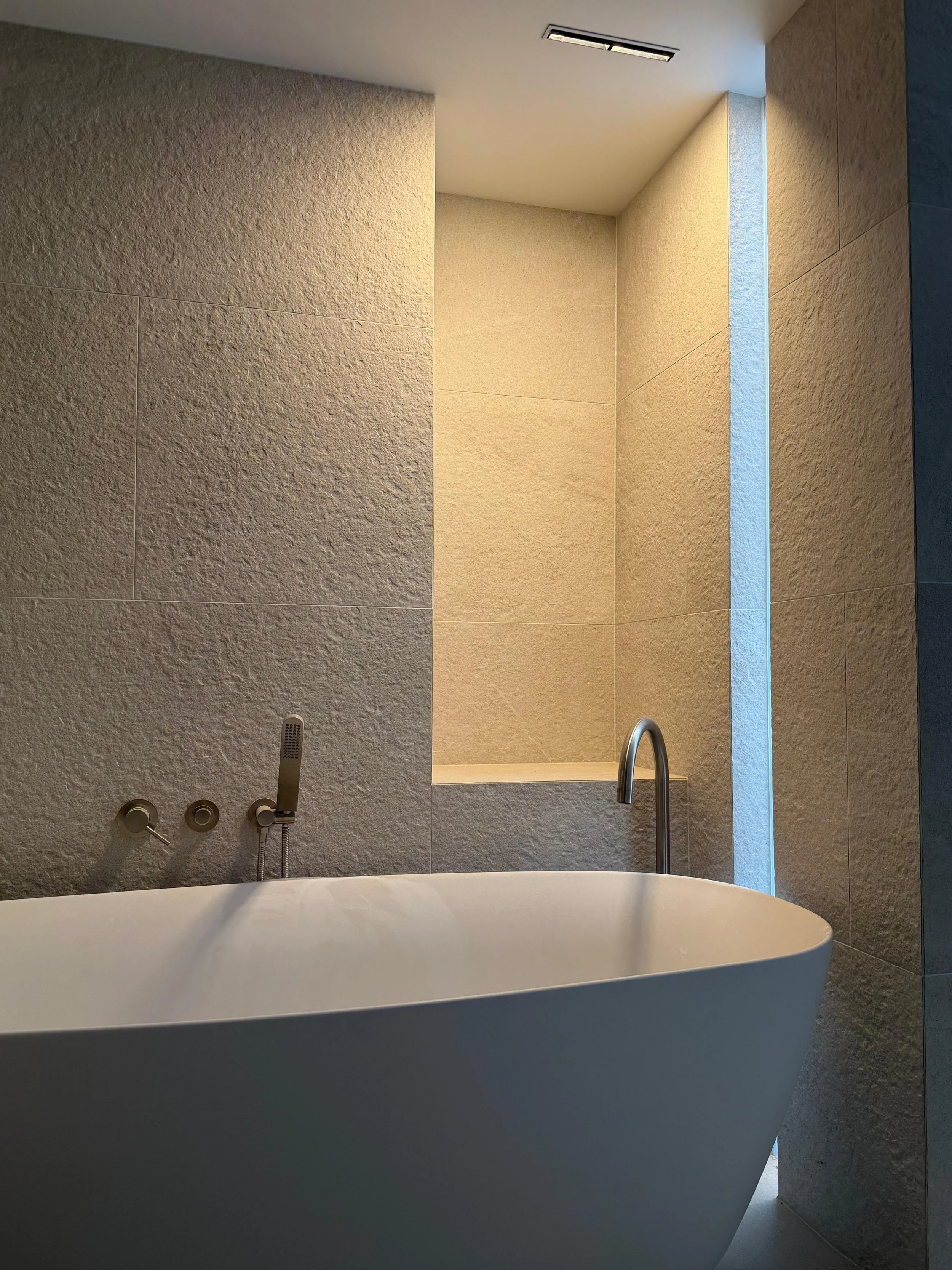 Modern freestanding bathtub in a minimalist bathroom with textured beige walls, a sleek faucet, and a small niche with lighting.