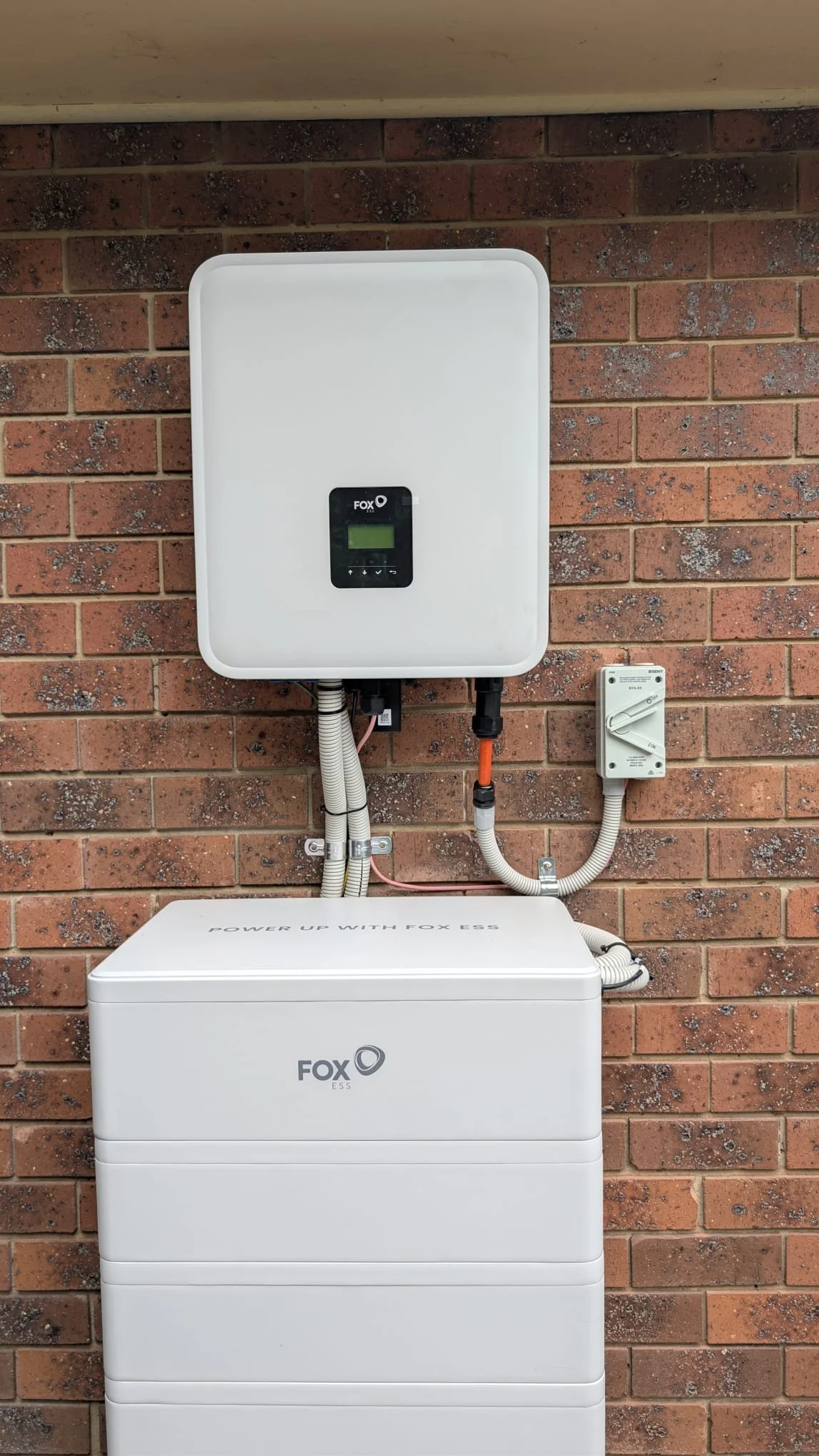 Photograph of a white solar energy system installation on a brick wall, showing an inverter, a power box, and connecting cables.