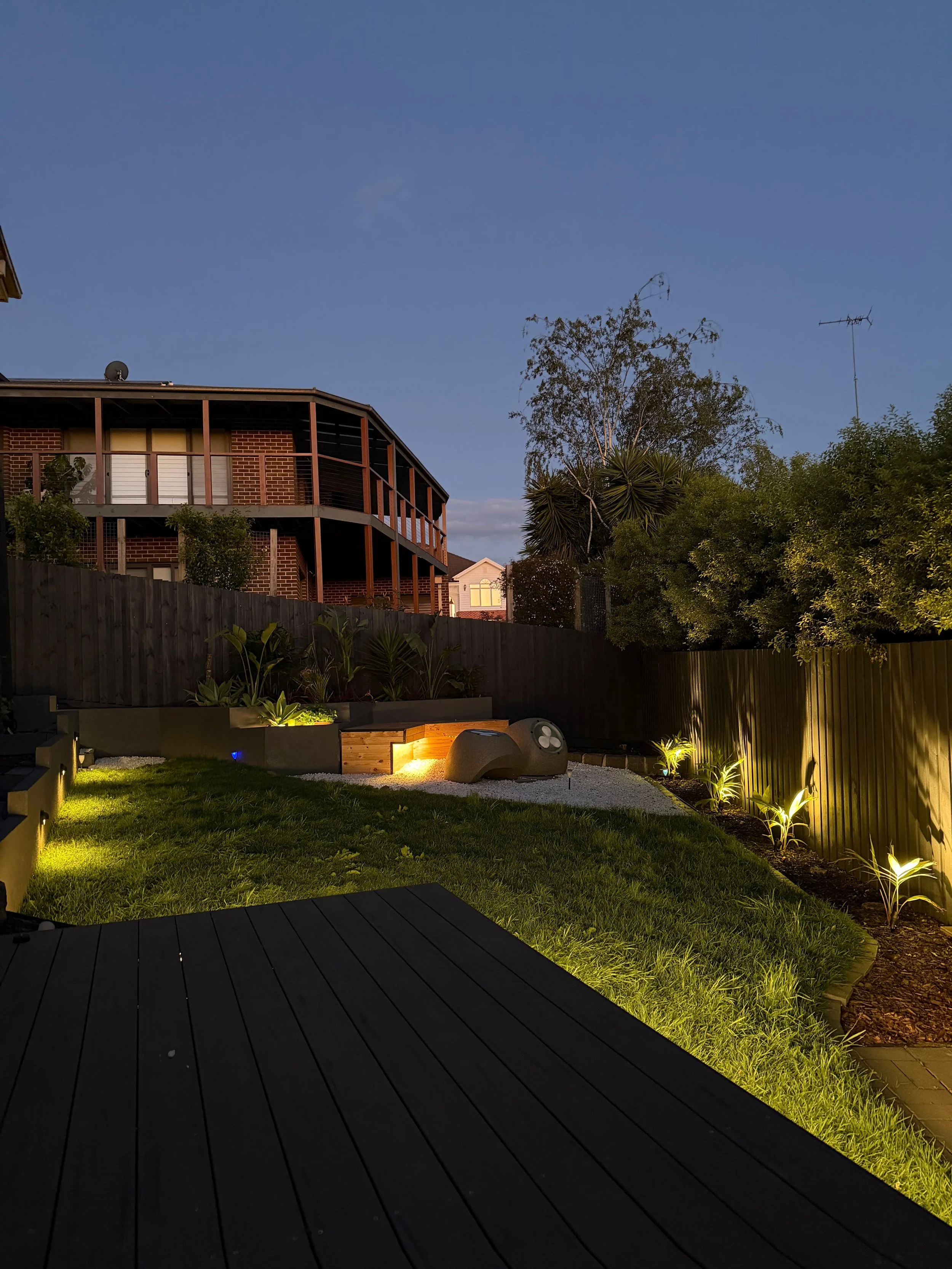 A backyard with a wooden deck, green grass, and landscaped plants illuminated by outdoor lighting at dusk. A modern sculpture or seating piece is visible on the lawn, with a dark fence and trees in the background.