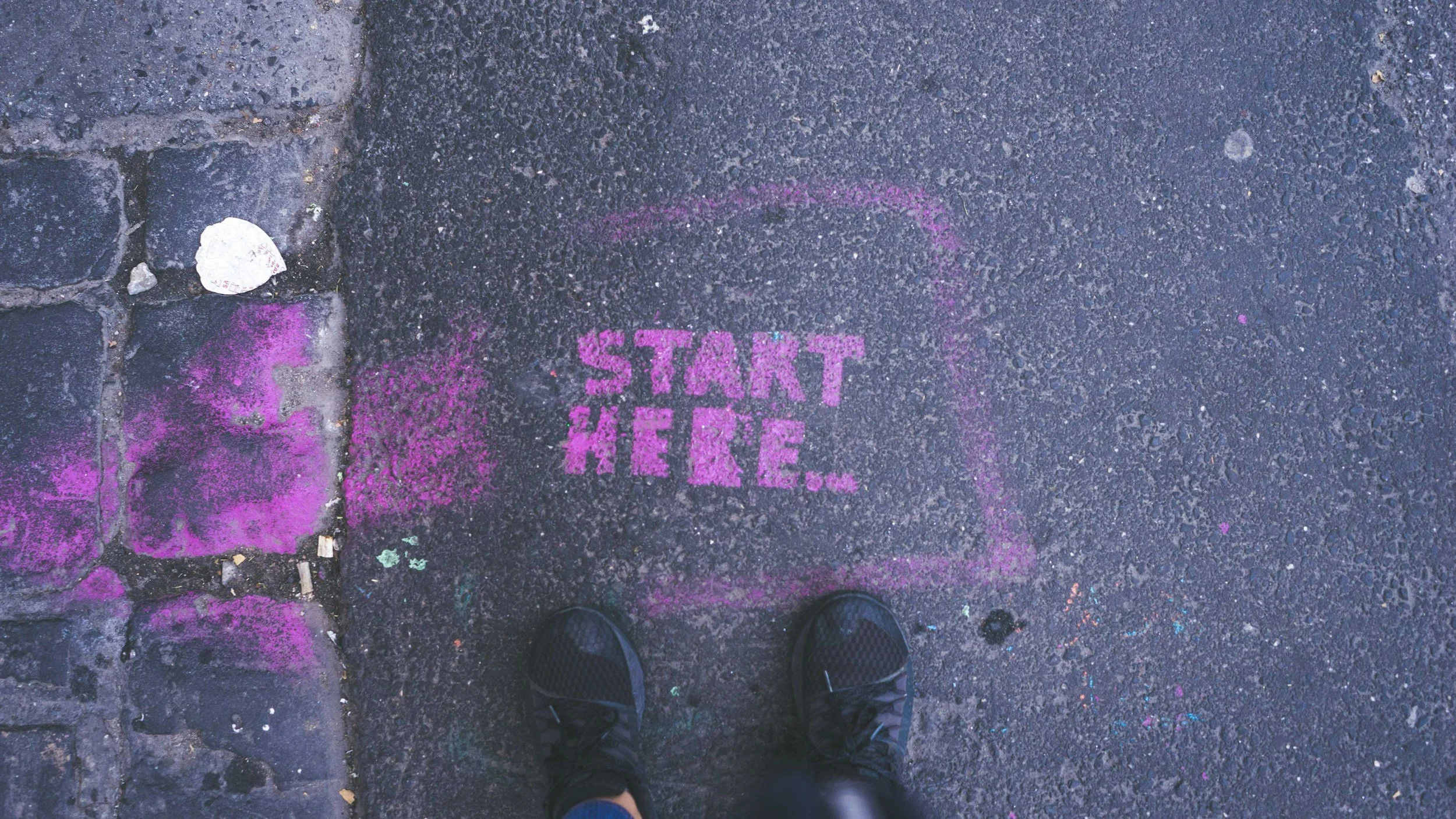Street with pink chalk stencil art that reads 'START HERE' on the pavement, with a person wearing black shoes standing above it.