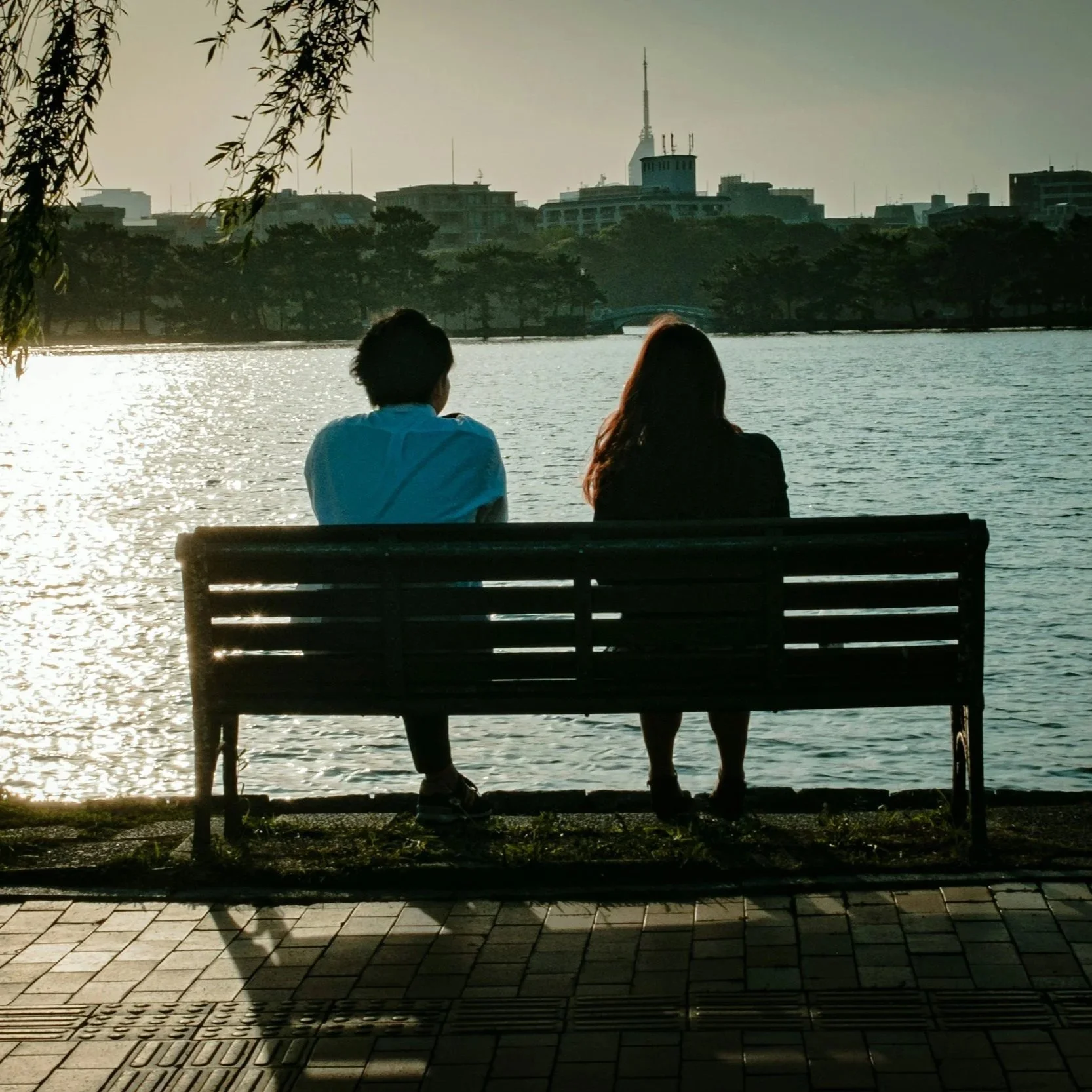 A man and woman sitting on a bench by a lake, watching the sunset with a city skyline and tall building in the background.