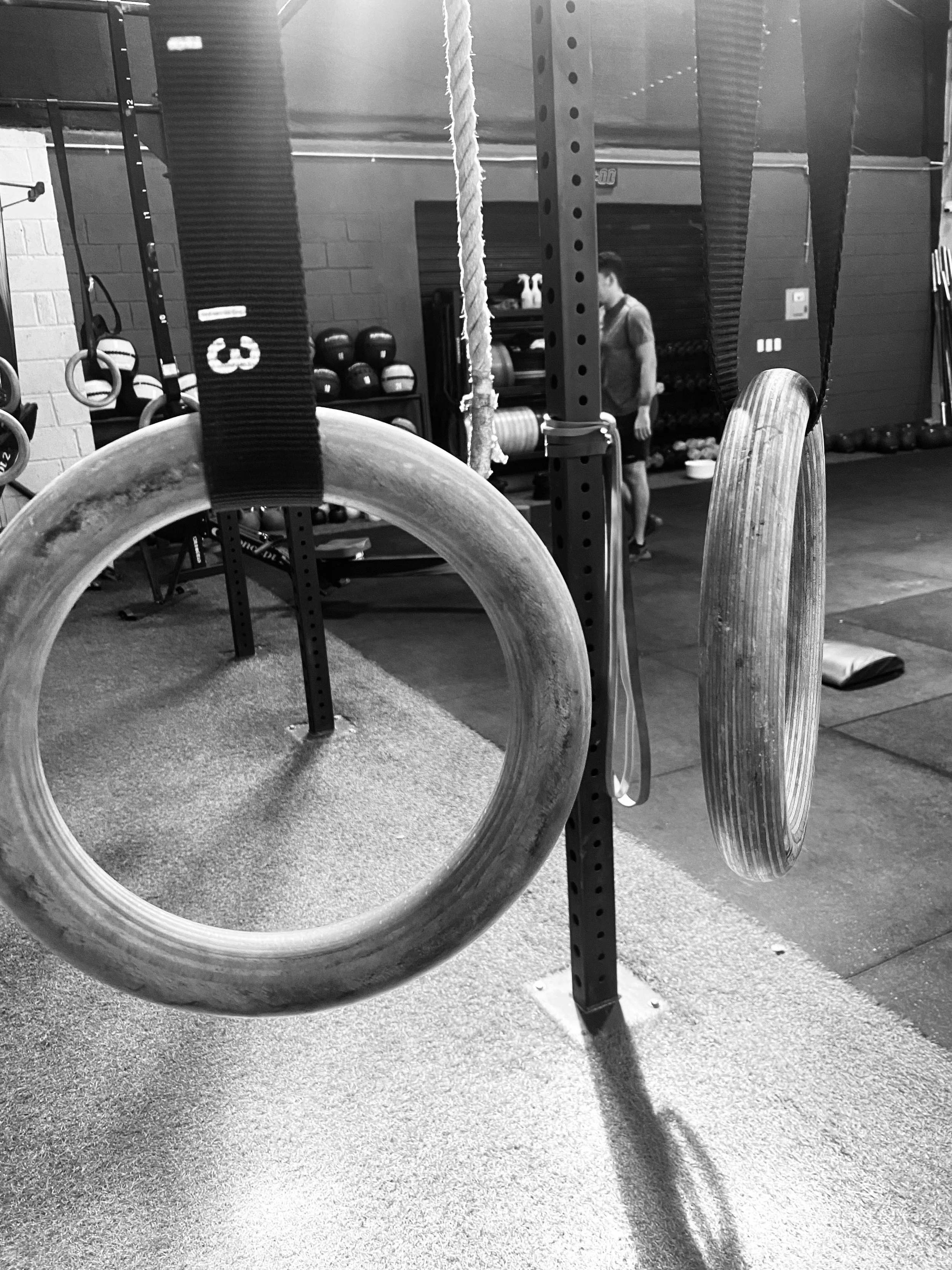 Black and white photo of gym equipment with two gymnastic rings hanging from straps in the foreground and a person standing in the background near a rack of kettlebells.