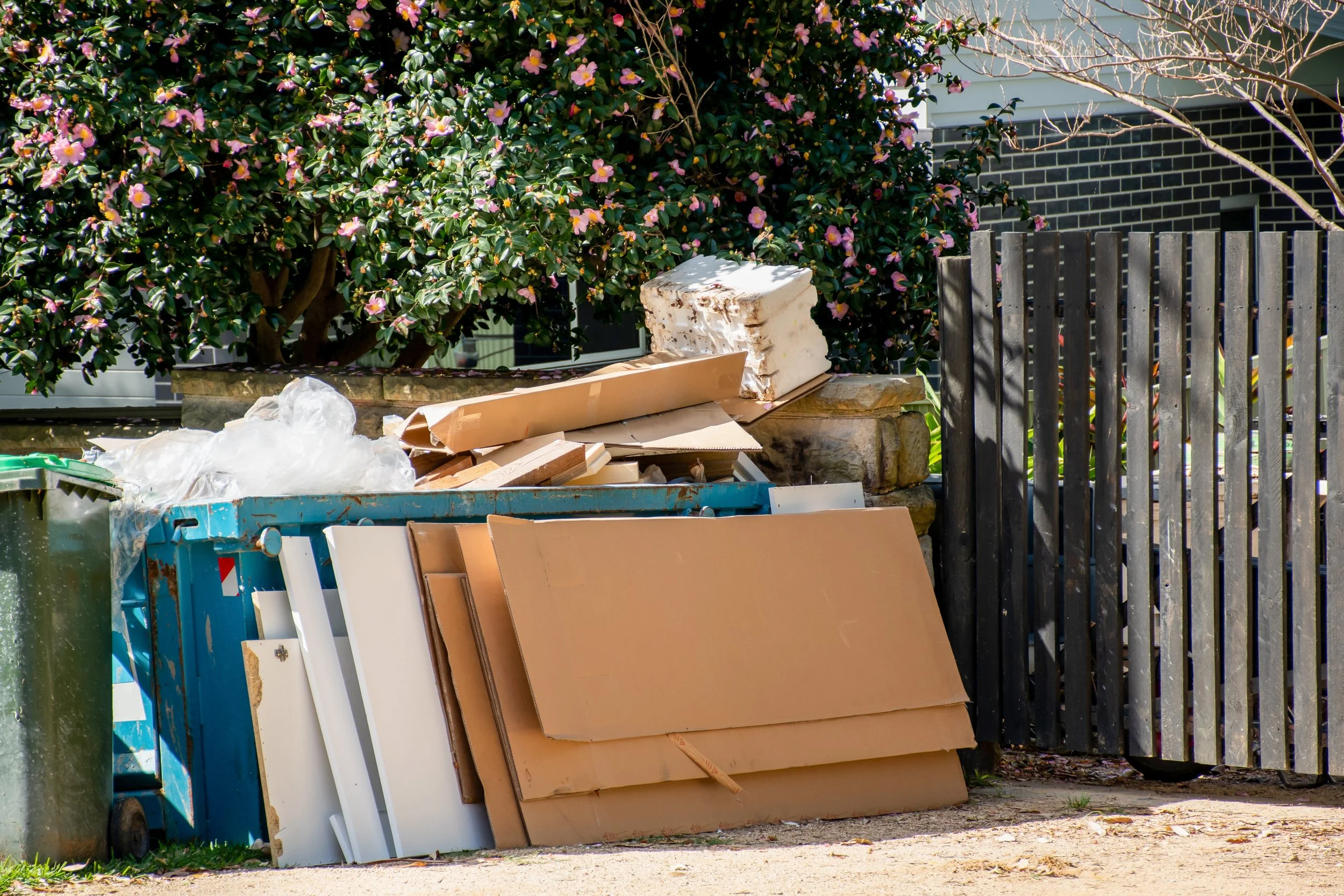 Overflowing trash and cardboard boxes outside near a bush and wooden fence.