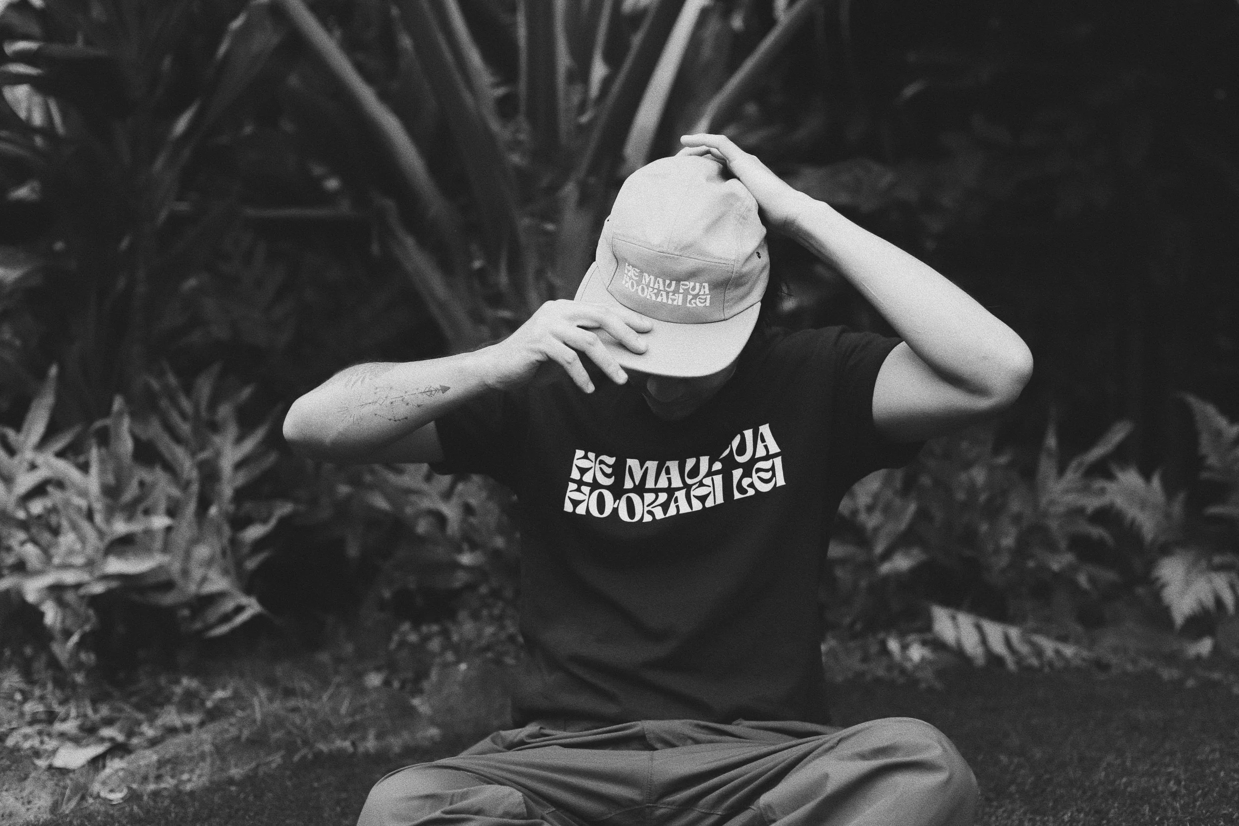 A person sitting outdoors, adjusting a cap with one hand and holding the brim with the other. The person is wearing a T-shirt and pants, with a backdrop of foliage.