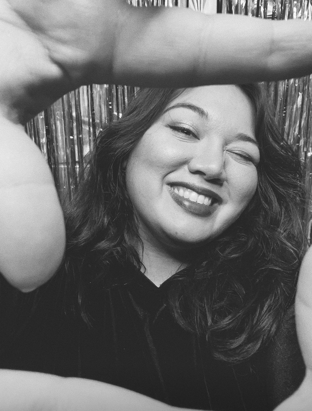 A black-and-white photo of a woman with curly hair smiling and winking, with her arm raised above her head, in front of a shiny, textured background.