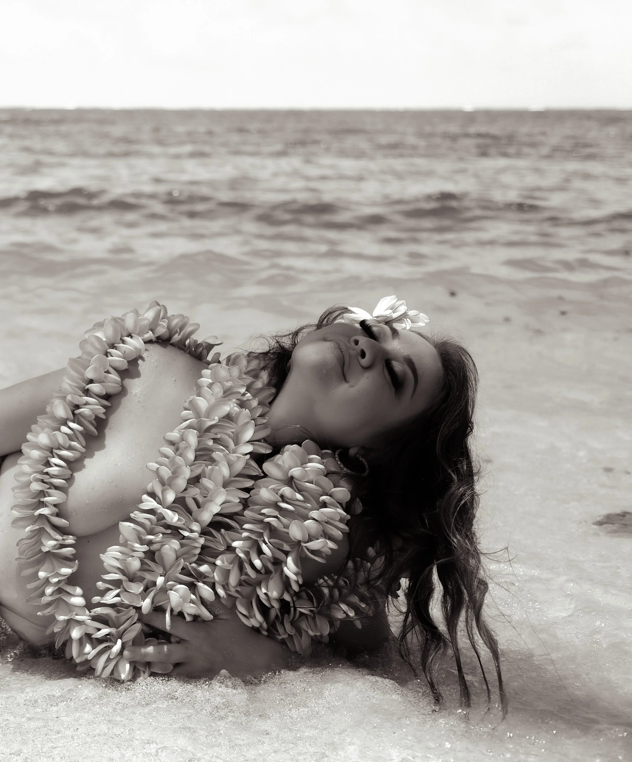 A woman wearing a floral lei and headpiece relaxing on the beach with her eyes closed and water around her.