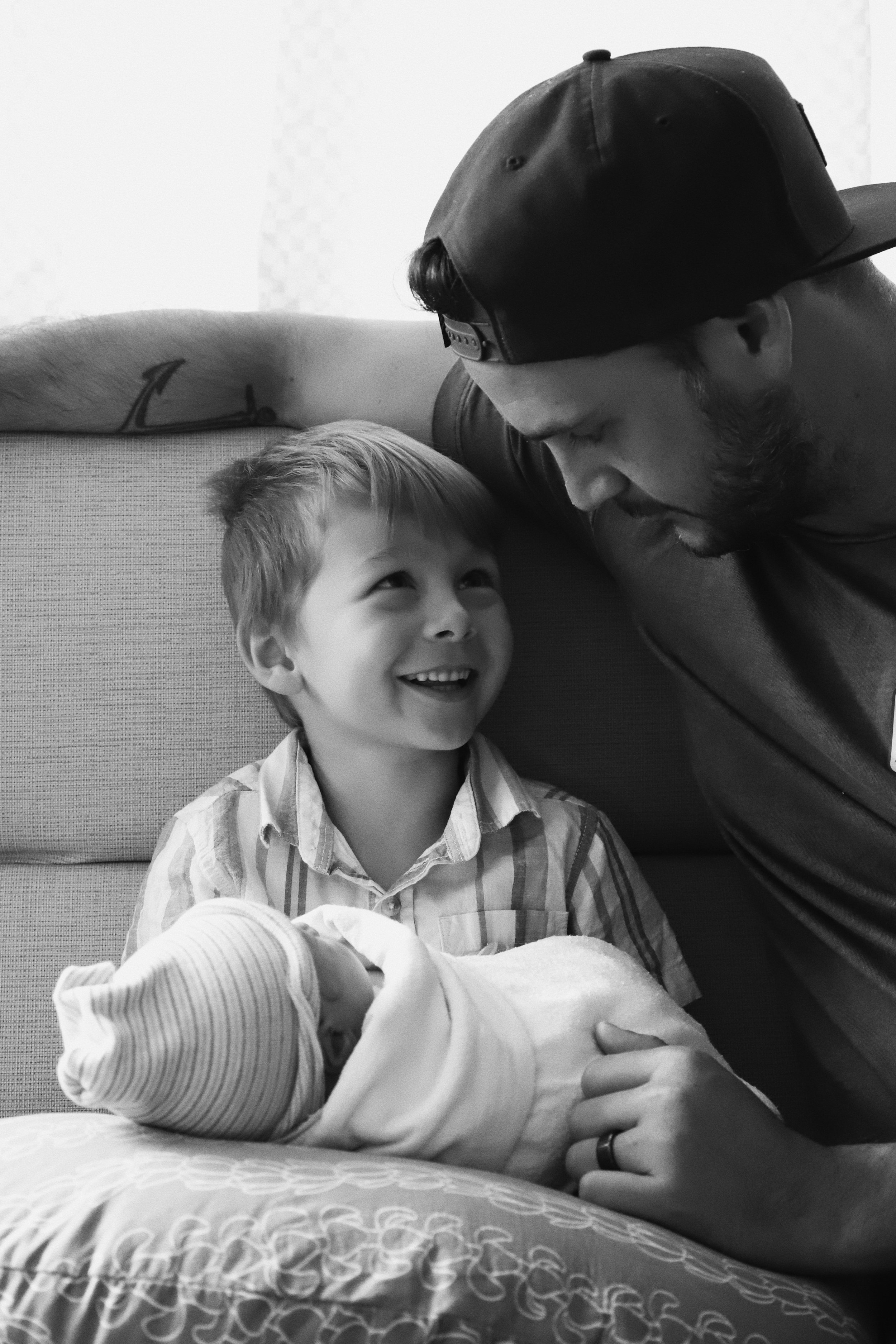 A young boy with a big smile looking at a man, likely his father, who is leaning over him with his forehead touching the boy's forehead. The boy is holding a newborn baby wrapped in a blanket, lying on a bed. The man, boy, and baby are in a close, in