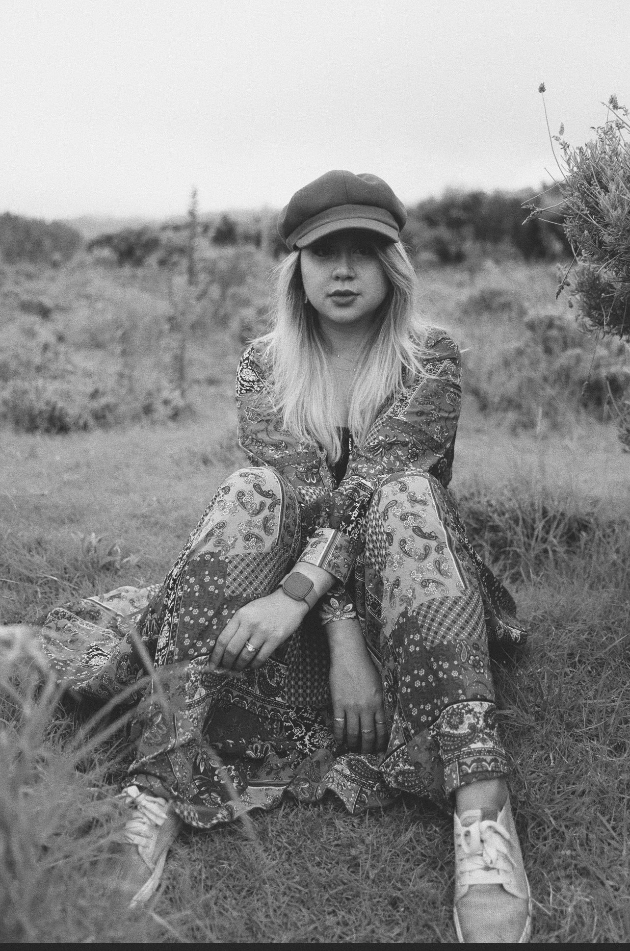 A young woman with long blonde hair and a hat sitting on grass outdoors. She is wearing patterned clothing and sneakers, with a neutral expression, in a black and white photograph.