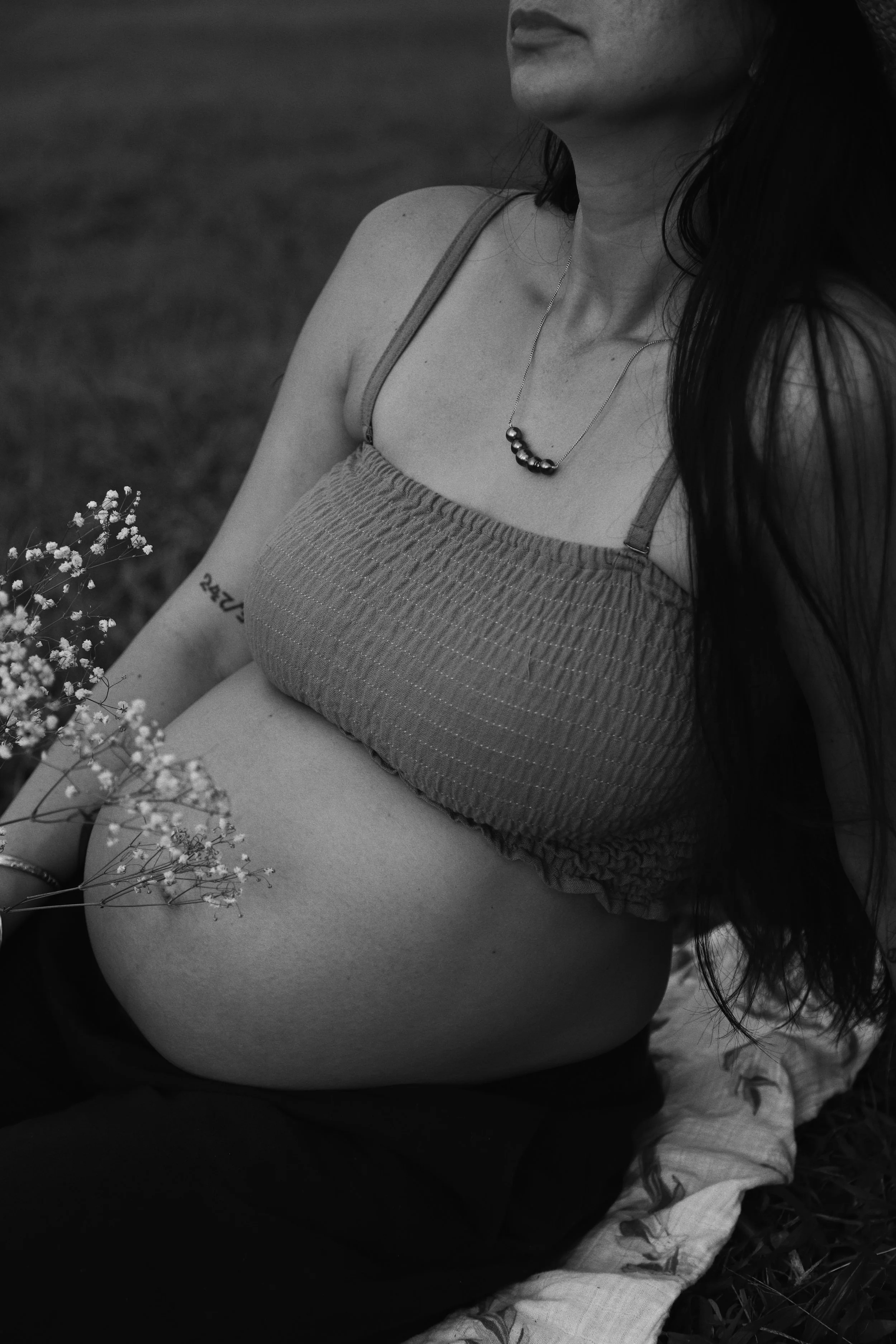 A black and white photo of a pregnant woman sitting outdoors on a blanket, holding flowers, with long hair, a necklace, and a tattoo on her arm.