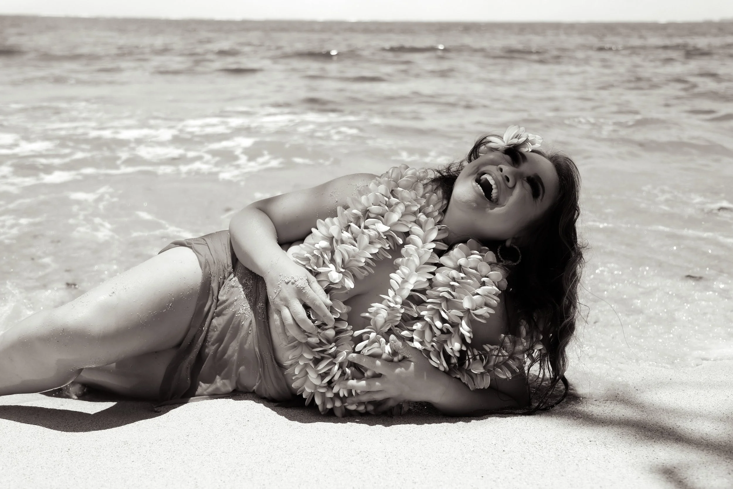 Woman lying on the beach, laughing, wearing a flower lei and flowers in her hair, with the ocean in the background.