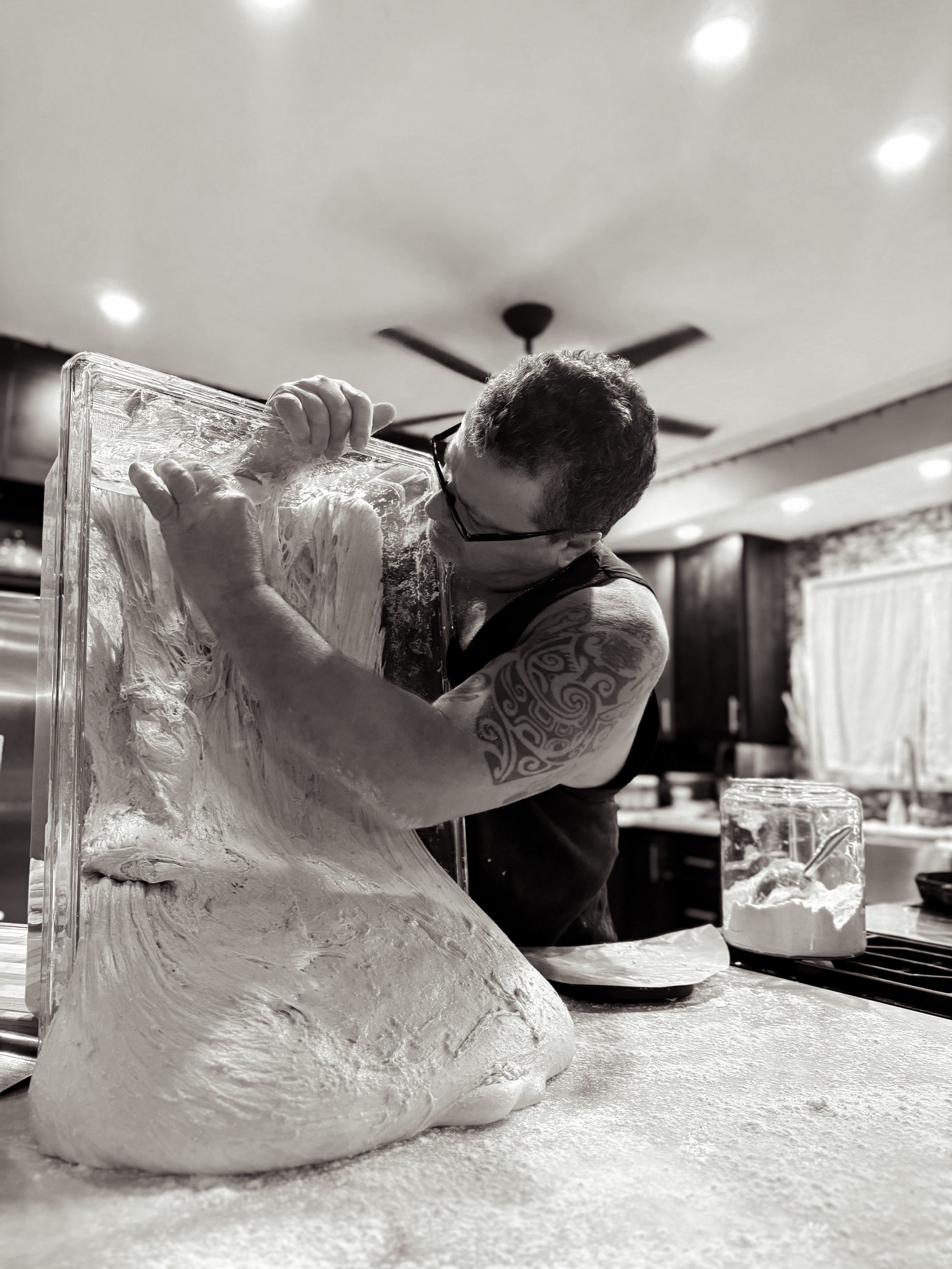 A man with tattoos on his arm and glasses is working with dough on a kitchen counter. He is handling a large, flat, transparent surface.