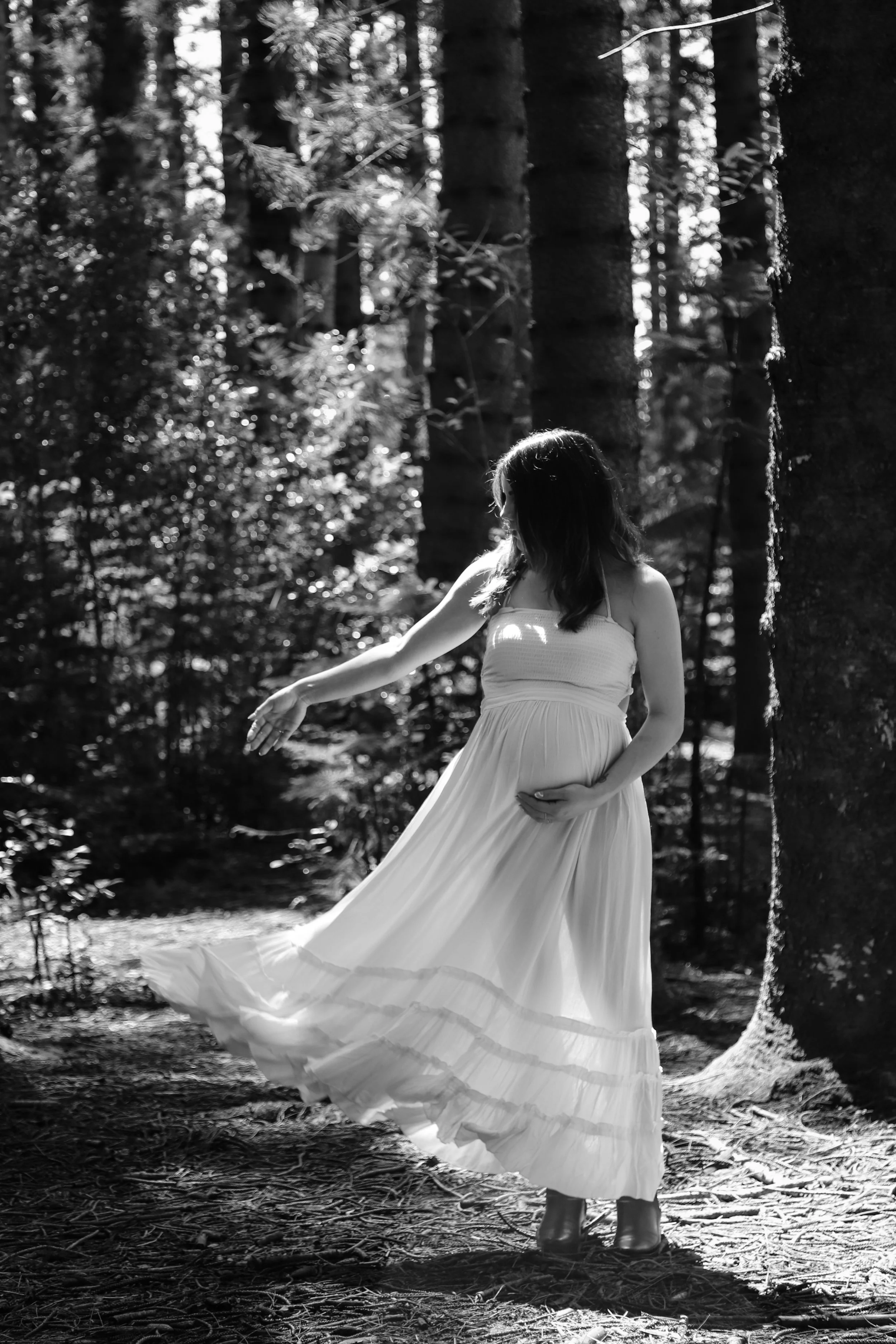 Black and white photo of a pregnant woman in a flowing dress standing in a wooded forest, with sunlight filtering through the trees.
