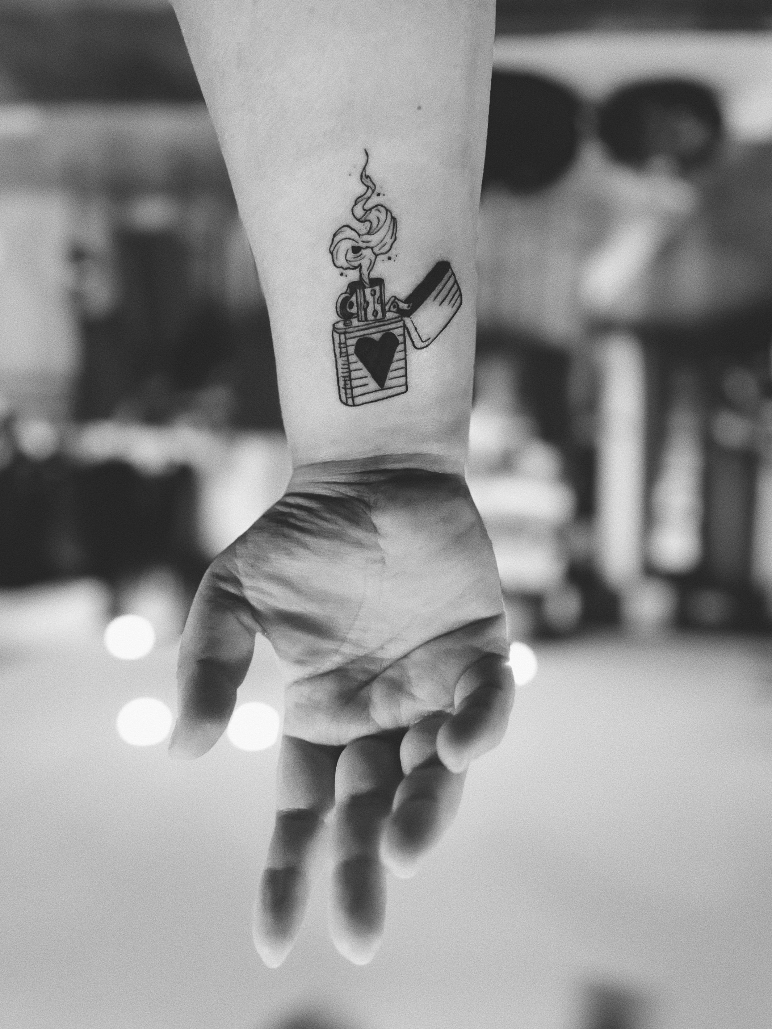 Black and white photo of a person's arm with a tattoo of an open lighter with a heart on it, and smoke rising from it. The person is holding their hand open with fingers relaxed.