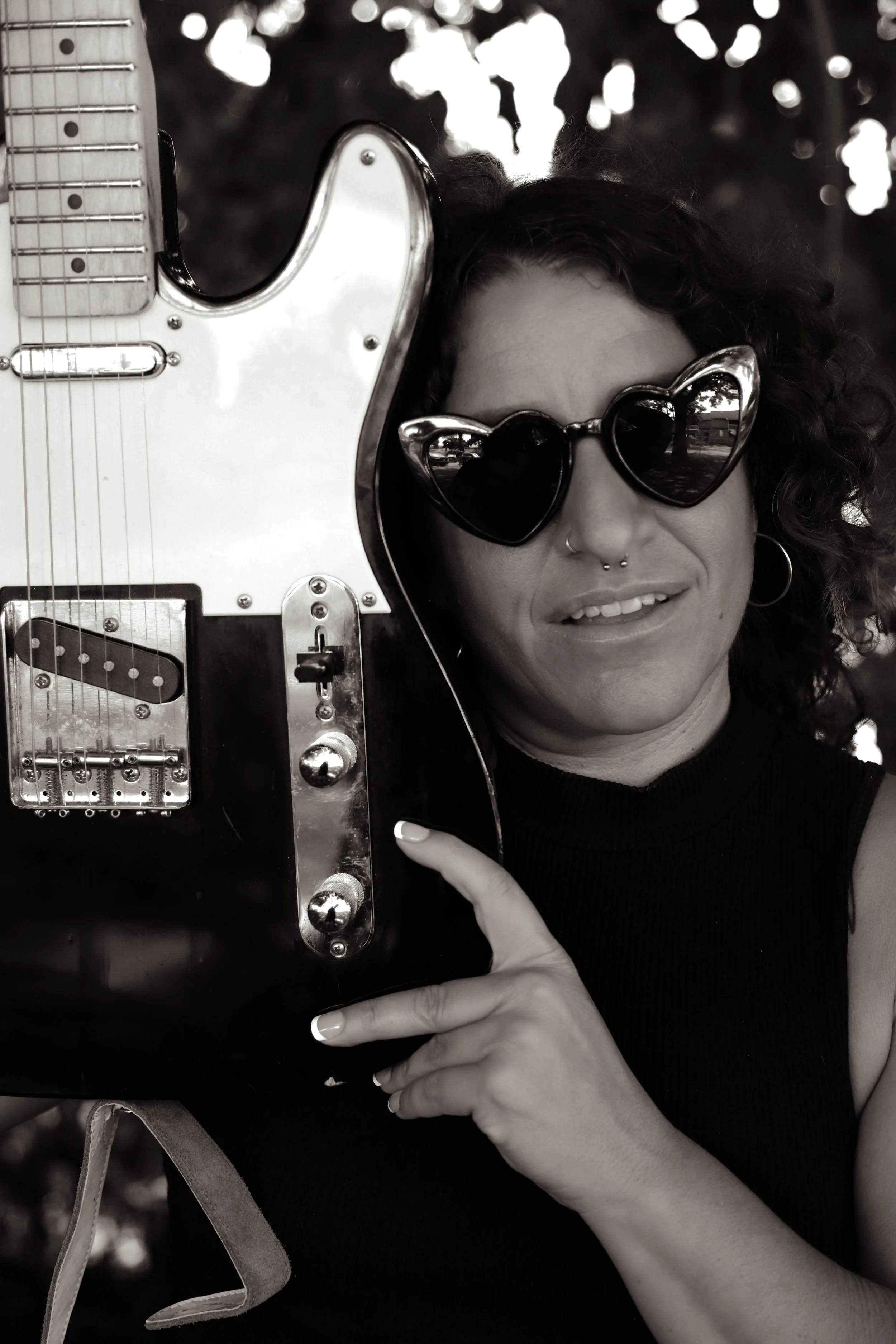Black and white photo of a woman with curly hair wearing heart-shaped sunglasses, holding a guitar near her face, and smiling.
