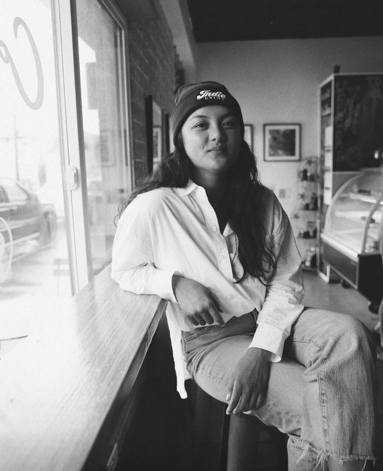 A young woman with long dark hair sitting on a stool by a large window, wearing a beanie, a white shirt, and jeans, in a cafe with framed pictures on the wall and a display case in the background, in black and white.