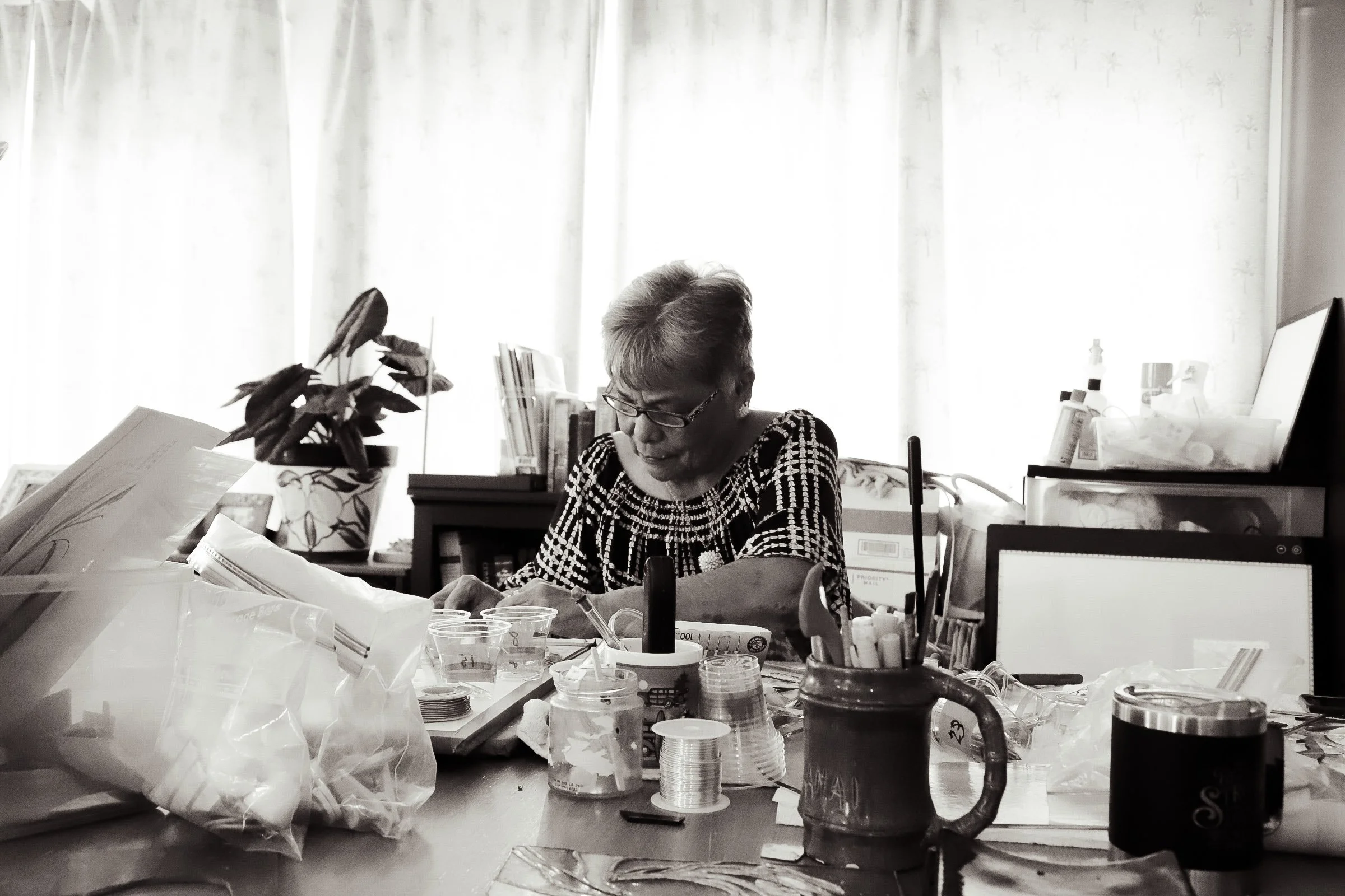 A woman sitting at a cluttered desk filled with papers, jars, a mug, and other office supplies, working or reading amidst a bright window with curtains in the background.